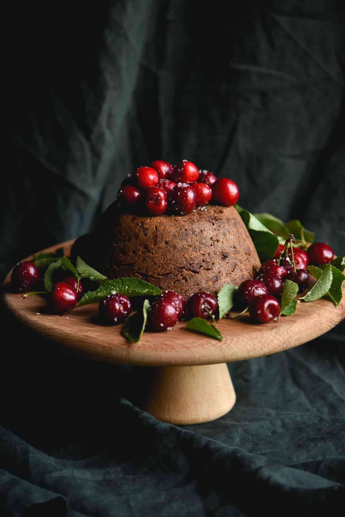 A side on view of a fruitless Christmas pudding topped with cherries against a green backdrop
