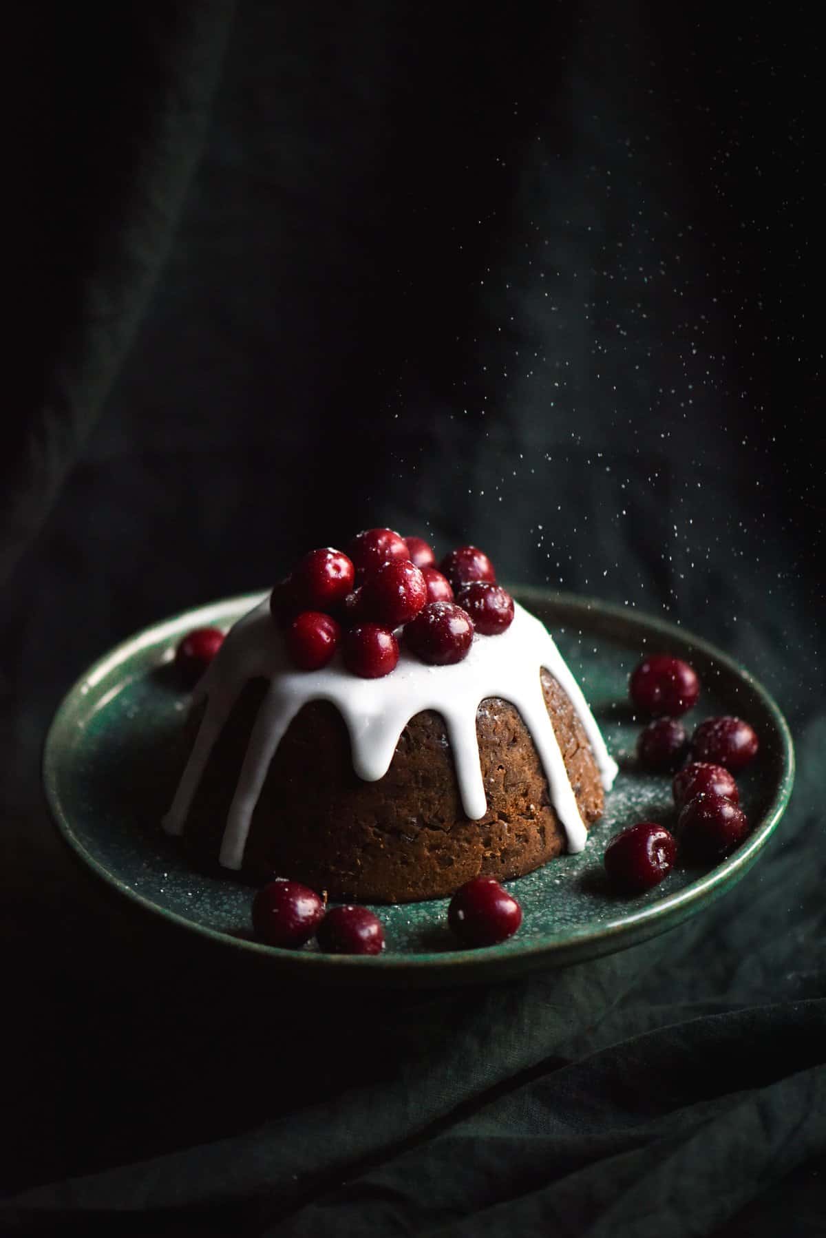 A side on photo of a fruitless, gluten free Christmas pudding, adorned with a white icing and topped with cherries
