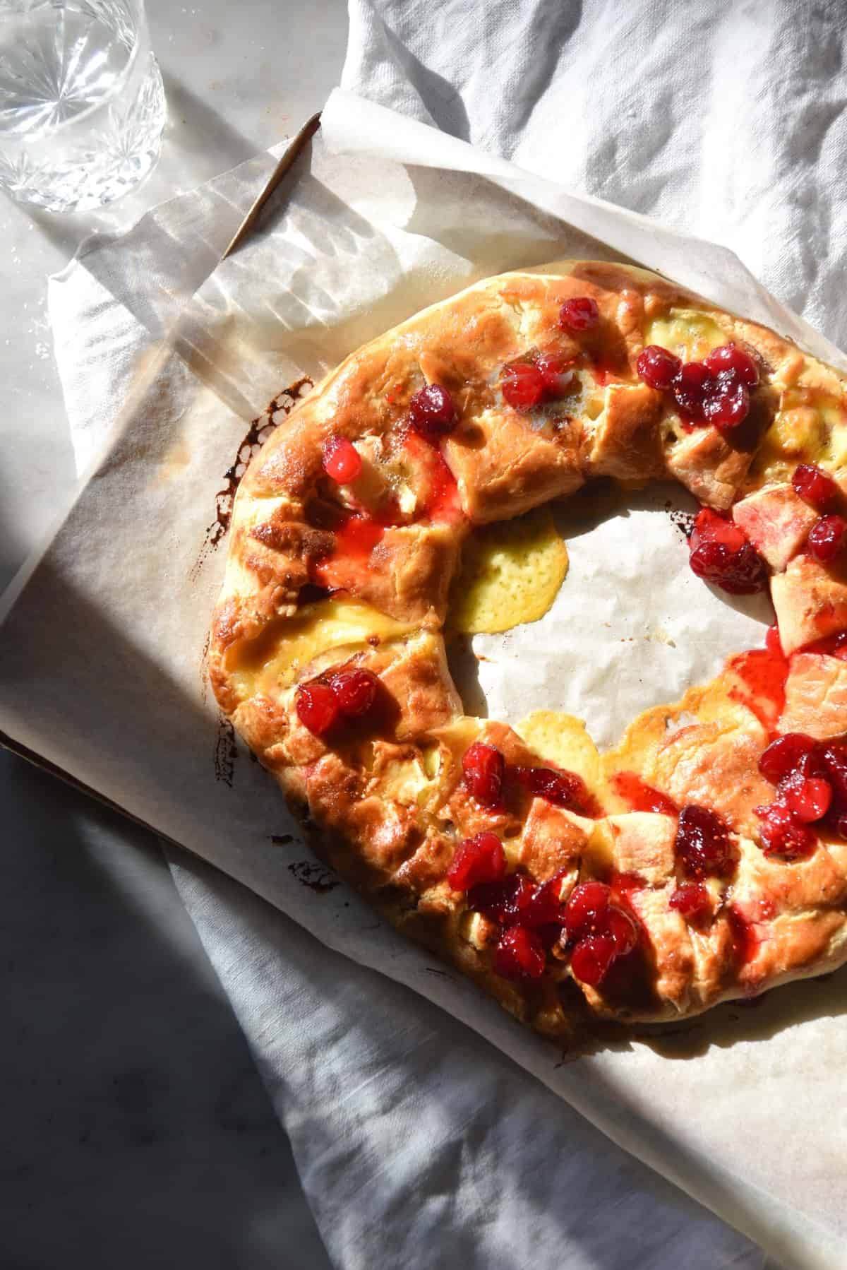 An aerial sunlit image of a gluten free brie and cranberry wreath on a lined baking tray on a white linen tablecloth