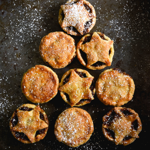 Gluten free, fruit free mince pies arranged in the shape of a Christmas tree on a dark steel backdrop. Some of the mince pies have full pastry tops, and some have cut out star decorations.