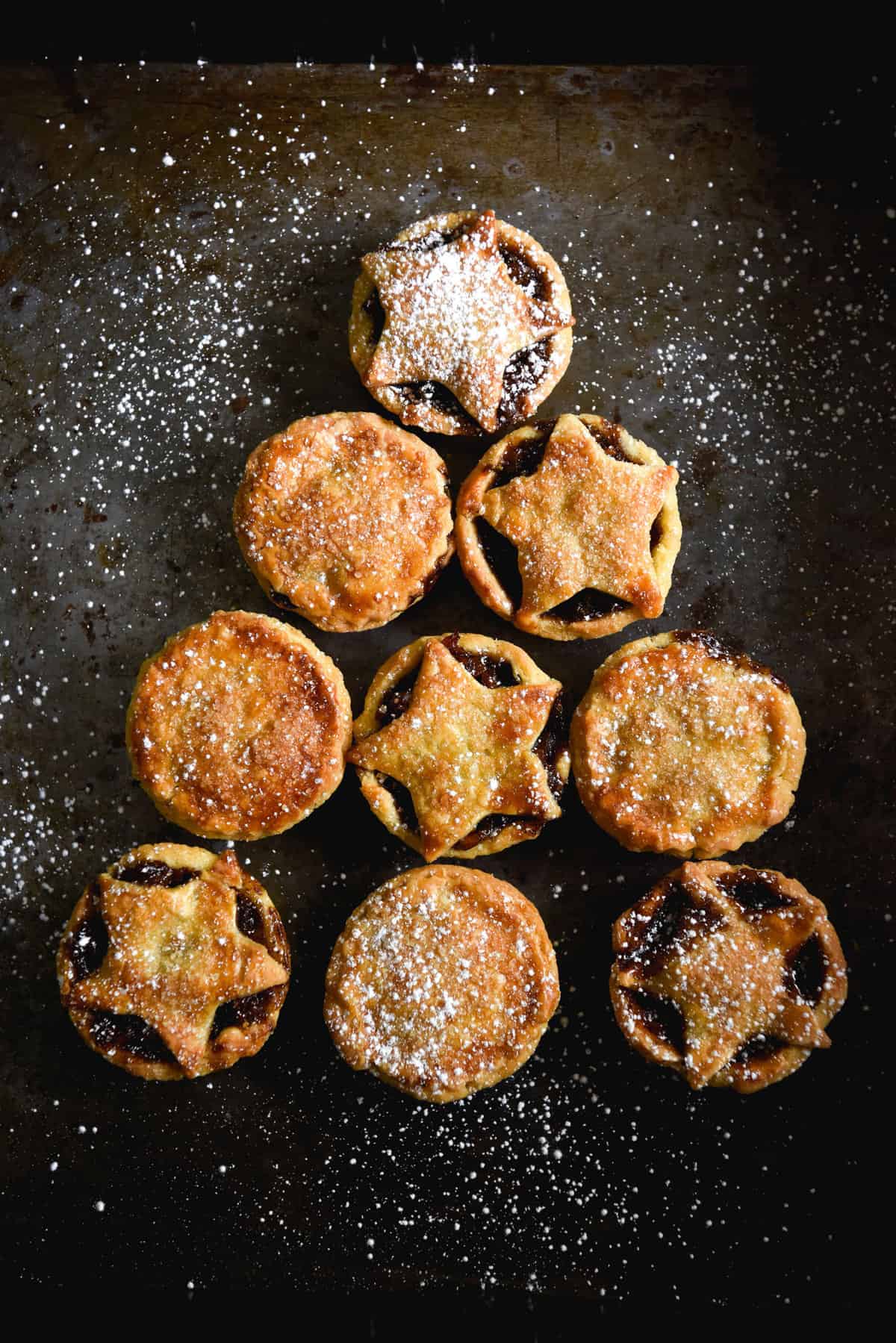 Gluten free, fruit free mince pies arranged in the shape of a Christmas tree on a dark steel backdrop. Some of the mince pies have full pastry tops, and some have cut out star decorations.
