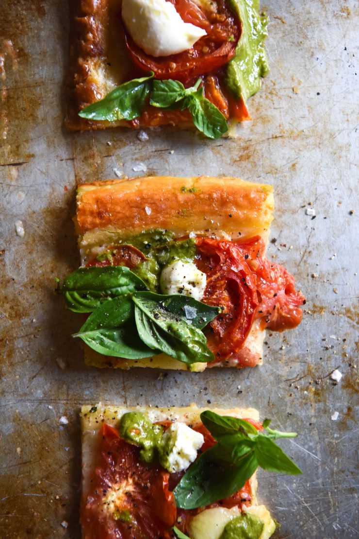 Squares of a gluten free tomato and pesto tart sit arranged in a line on a mottled sliver baking dish.