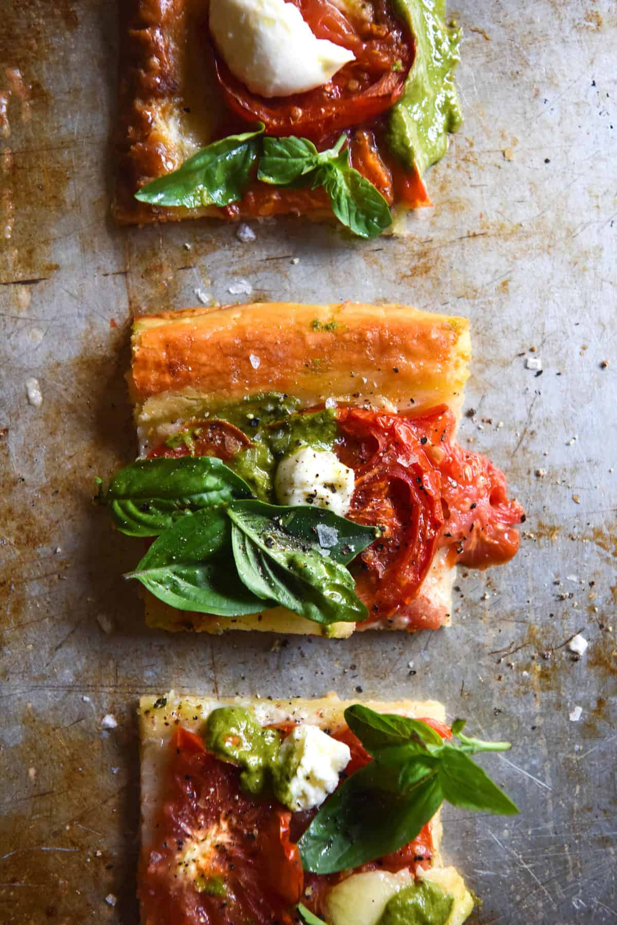 Squares of a gluten free tomato and pesto tart sit arranged in a line on a mottled sliver baking dish. 