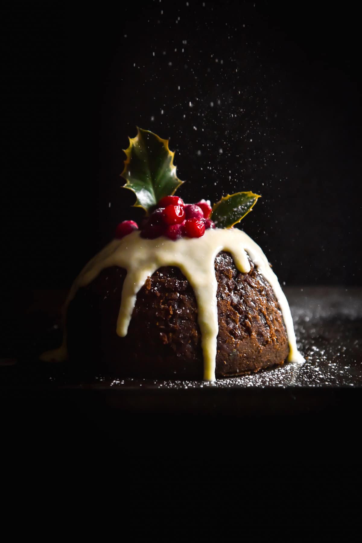A side on moody image of a gluten free Christmas pudding topped with icing, cranberries and holly leaves.  