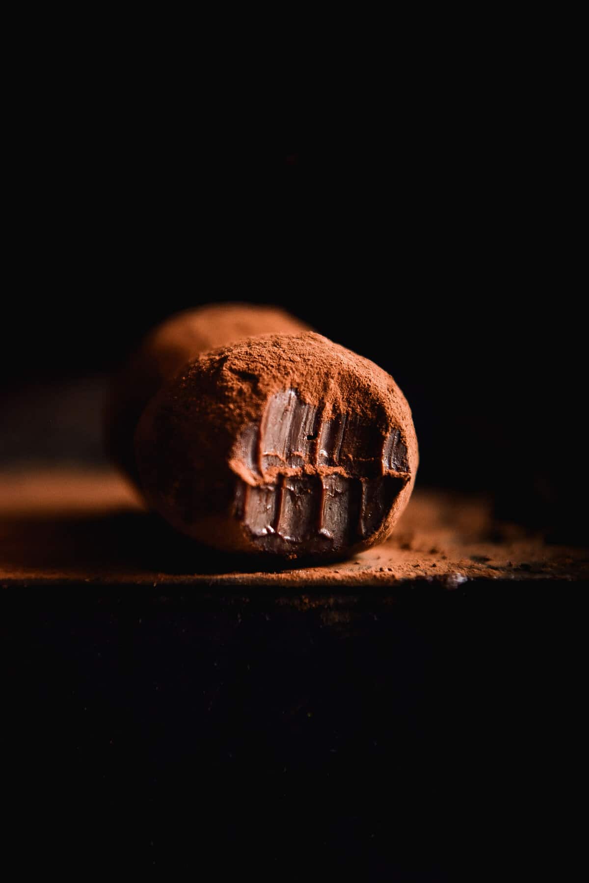 A moody side on image of a vegan Nutella truffle against a black backdrop. The truffle is coated with cocoa powder and has a bite mark taken out of the front of the truffle, exposing the rich chocolate inside.