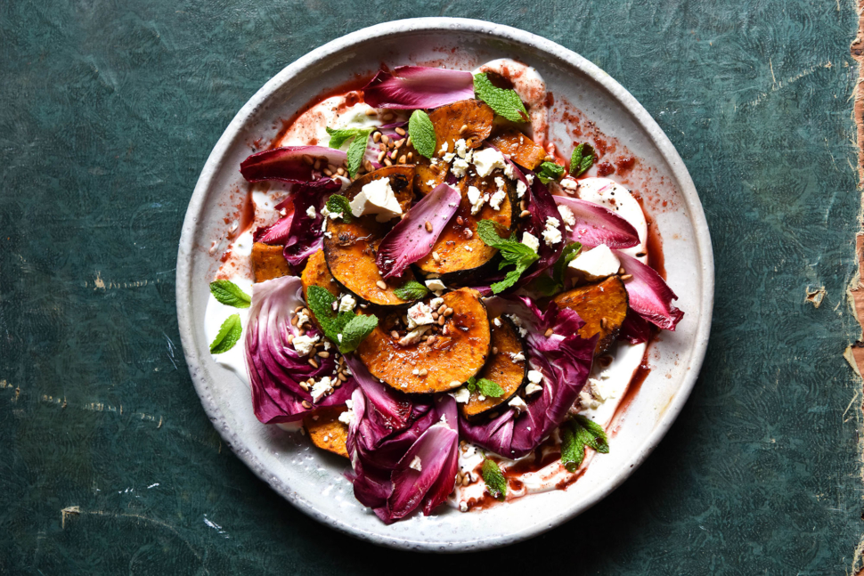 An aerial photo of a spiced pumpkin salad with radicchio, feta, pine nuts, mint and Christmas salad on a white ceramic serving platter atop an olive green tabletop. 
