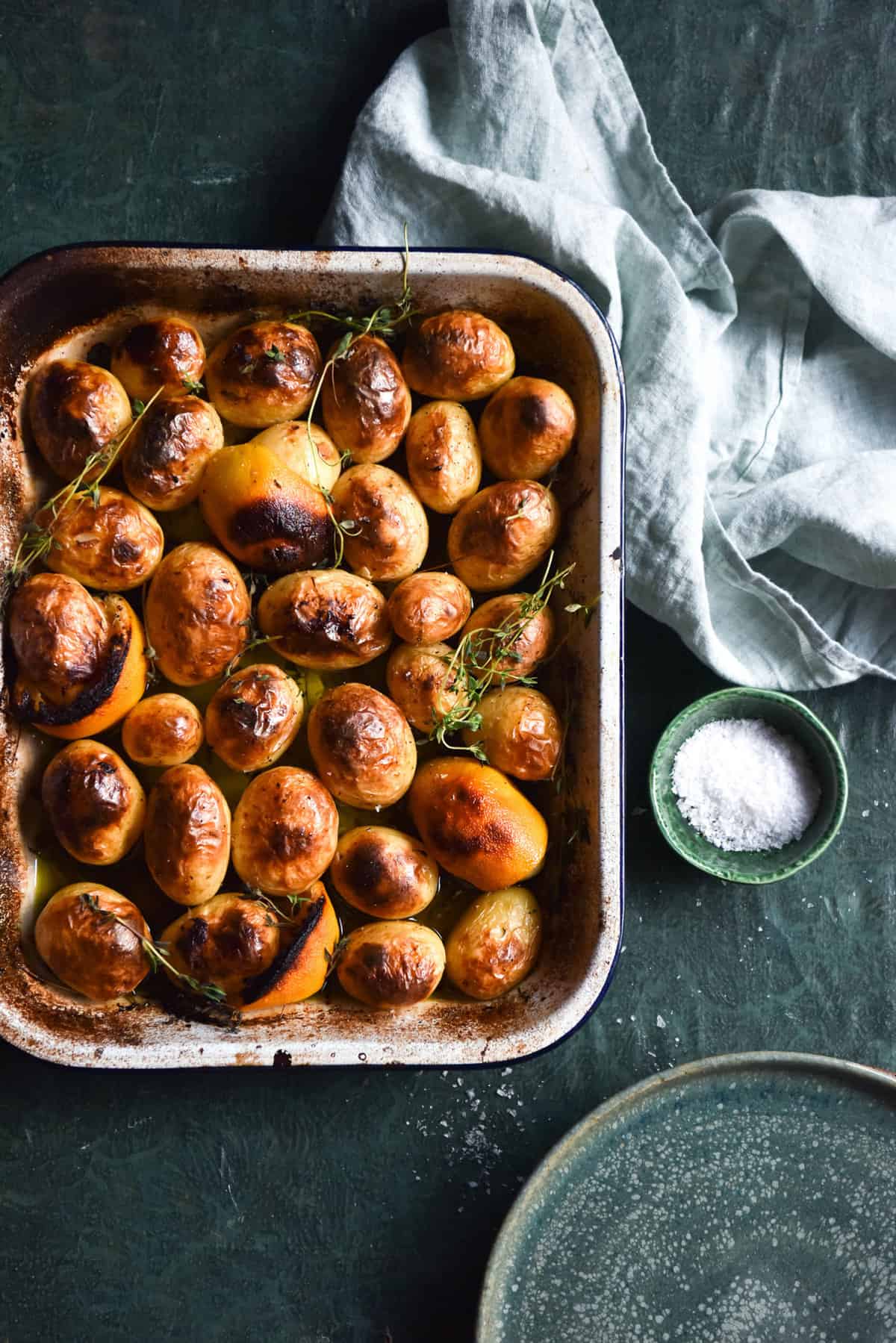 An aerial image of Greek lemon potatoes in a baking dish. The potatoes are golden brown, as are the lemons. The dish sits atop an olive green textured table and is surrounded by water glasses, extra plates and a tea towel.