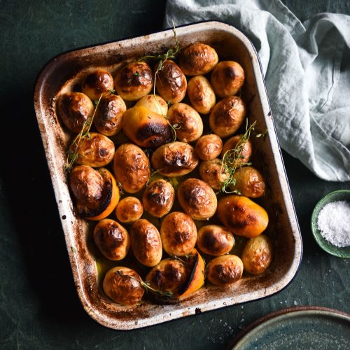 An aerial image of Greek lemon potatoes in a baking dish. The potatoes are golden brown, as are the lemons. The dish sits atop an olive green textured table and is surrounded by water glasses, extra plates and a tea towel.