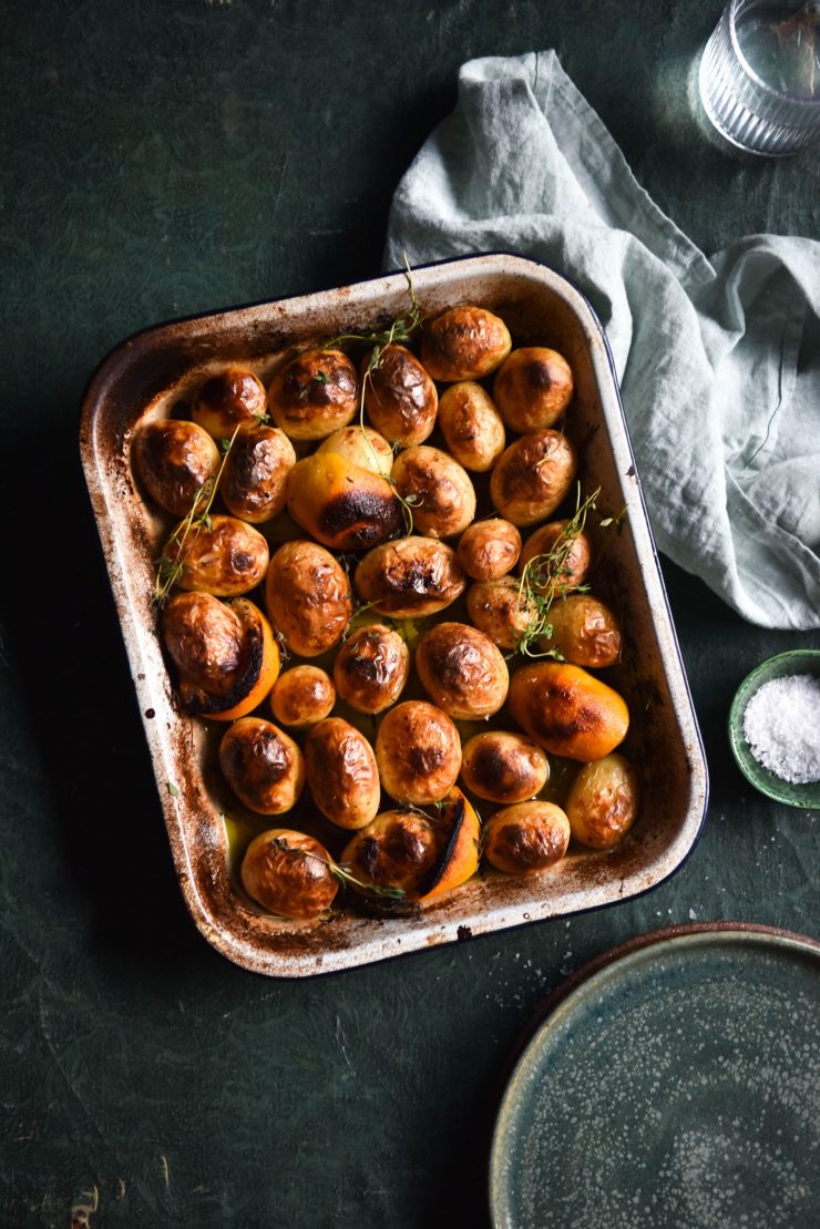 An aerial image of Greek lemon potatoes in a baking dish. The potatoes are golden brown, as are the lemons. The dish sits atop an olive green textured table and is surrounded by water glasses, extra plates and a tea towel.