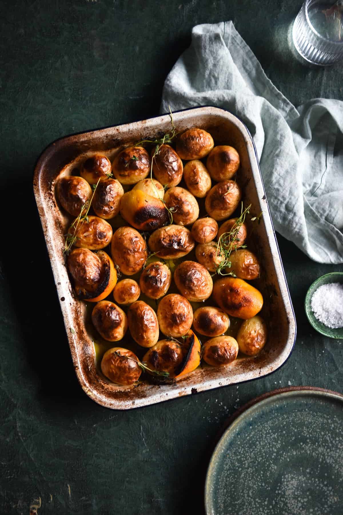 Lemon roasted potatoes in a white baking dish atop a dark green backdrop. The baking dish is surrounded by a sage green linen cloth, a green plate and a pinch bowl of salt