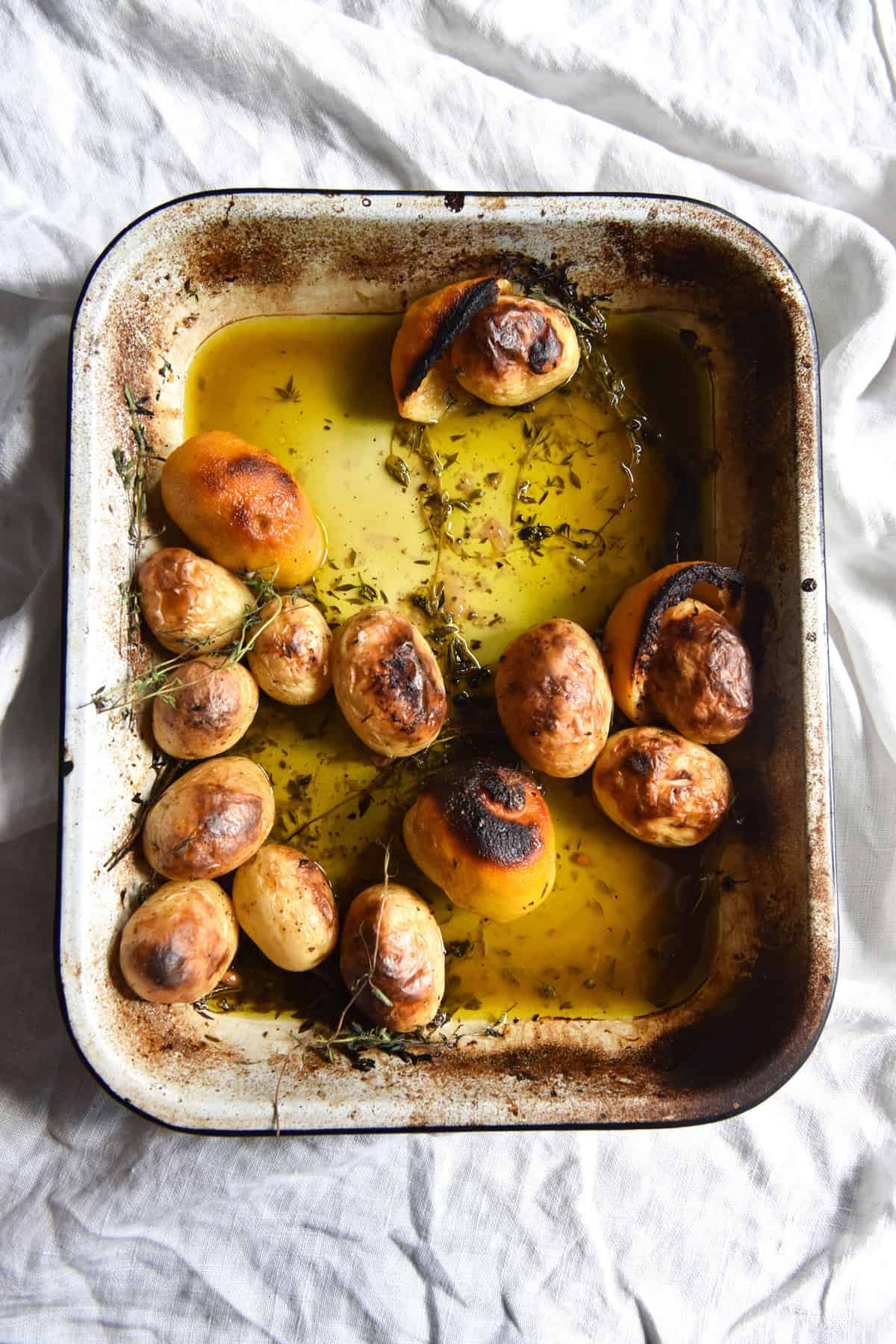 An aerial image of Greek lemon potatoes in a baking tray atop a white linen tablecloth. 