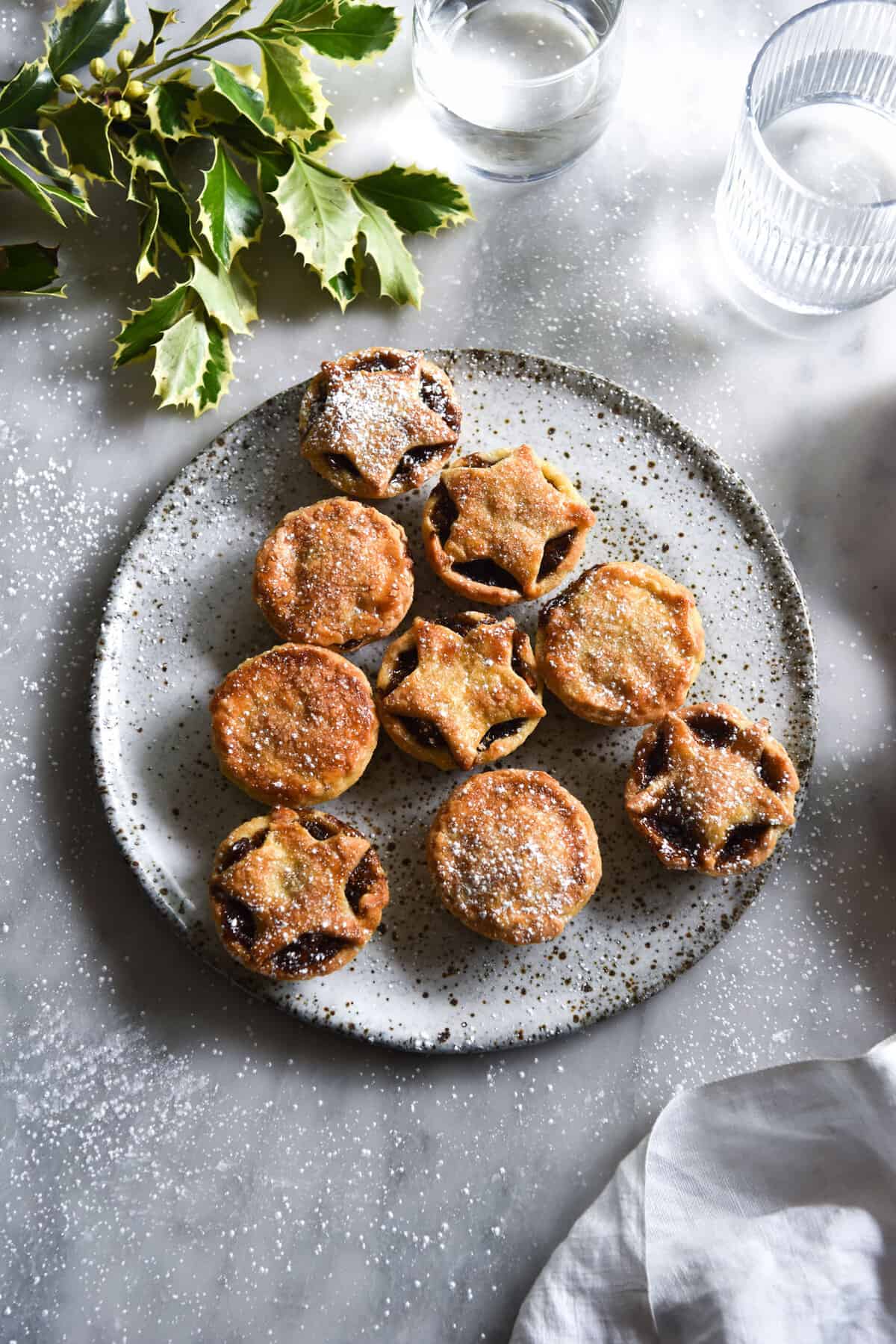 An aerial image of a white ceramic speckled plate topped with gluten-free fruit-free mince pies arranged in a Christmas tree shape. The plate sits on a white marble table surrounded by water glasses and holly leaves. 