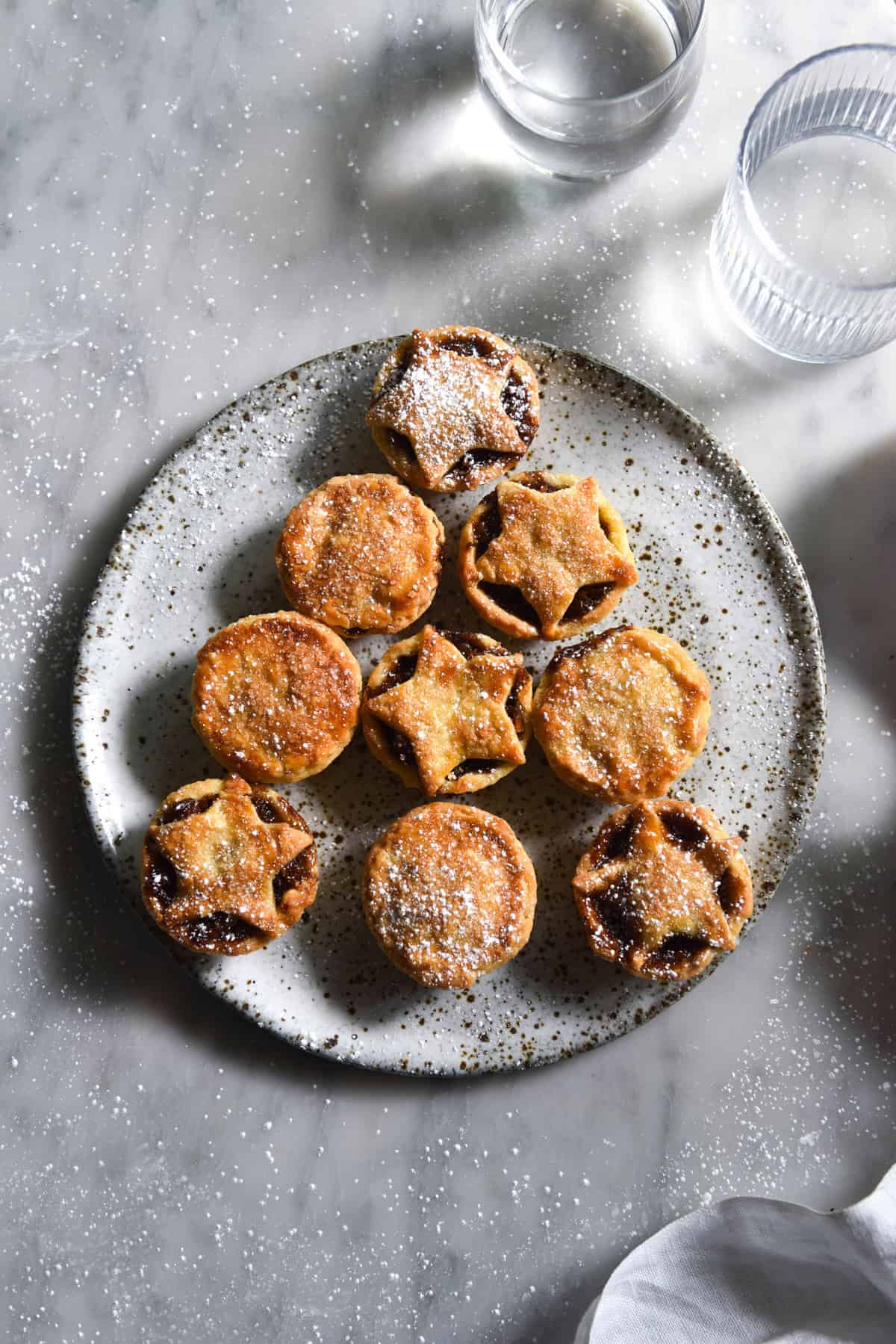 Gluten free, fruit free mince pies arranged in the shape of a Christmas tree on a white speckled ceramic plate atop a white marble table. Some of the mince pies have full pastry tops, and some have cut out star decorations. Glasses of water cast a light and shadow across the right hand side of the image