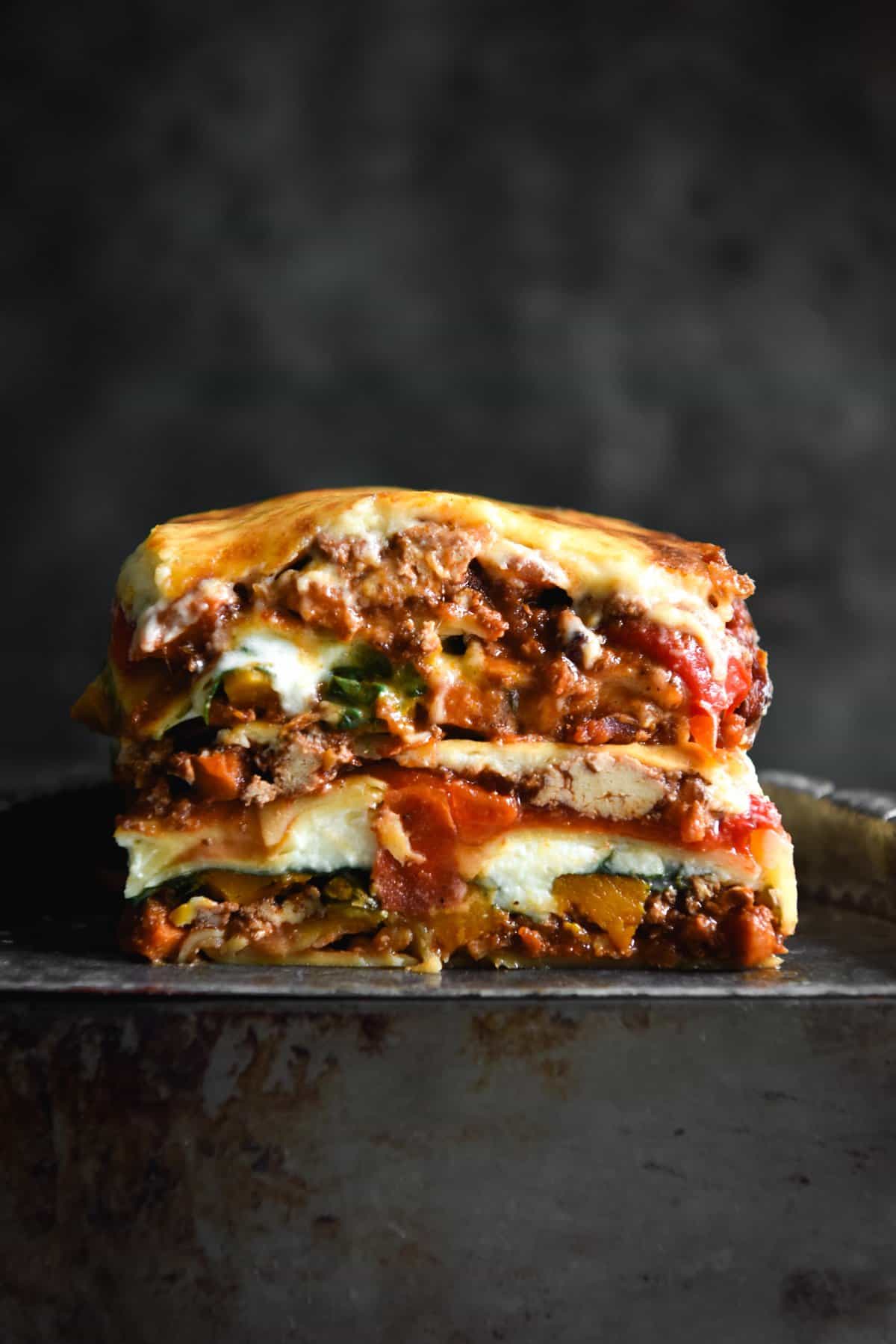 A side on macro image of two slices of gluten free vegetarian lasagne on a dark steel tray against a dark backdrop. 