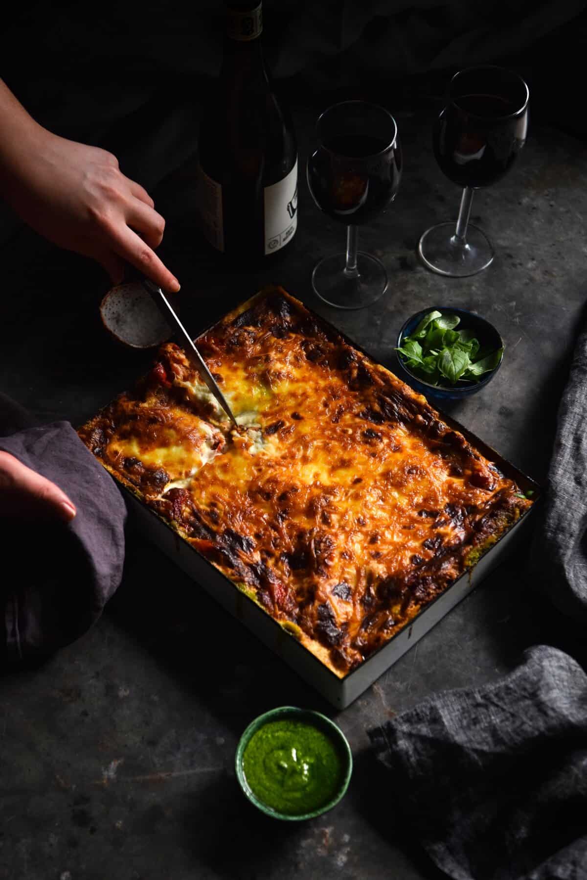 A moody image of a gluten free vegetarian and low FODMAP lasagne on a dark steel mottled backdrop. Hands emerge from the left side of the image to slice into the lasagne. It is surrounded by wine glasses and small pinch pots of herbs. 