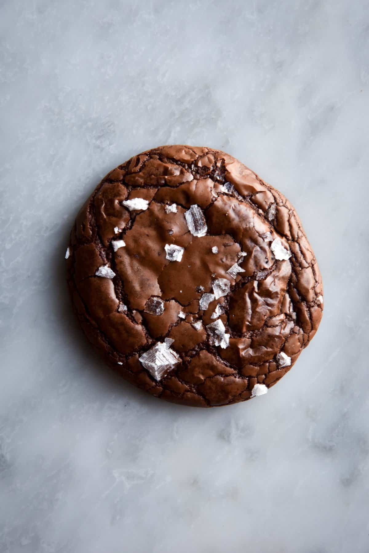 An aerial image of a gluten free brownie cookie on a white marble table. The cookie is crinkled and covered in sea salt flakes. 