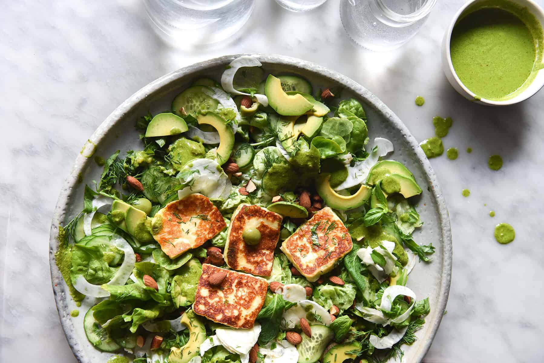 Green salad with haloumi, avocado and fennel topped with a FODMAP friendly almond pesto. It sits on a white ceramic serving platter on a white marble table. Glasses of water sit in the top of the image and cast pleasant light over the salad.