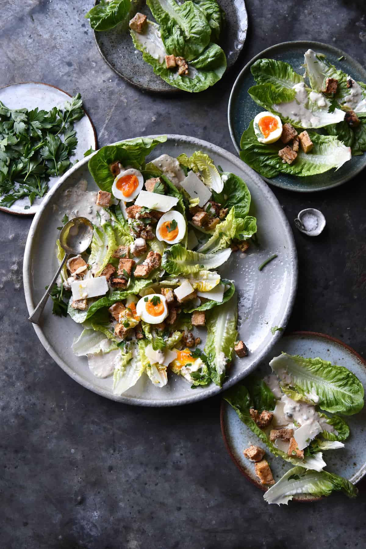 An aerial image of a white ceramic plate topped with vegetarian Caesar salad casually arranged on the plate. The central plate is surrounded by smaller plates also topped with salad. The scene is set on a dark blue steel mottled backdrop