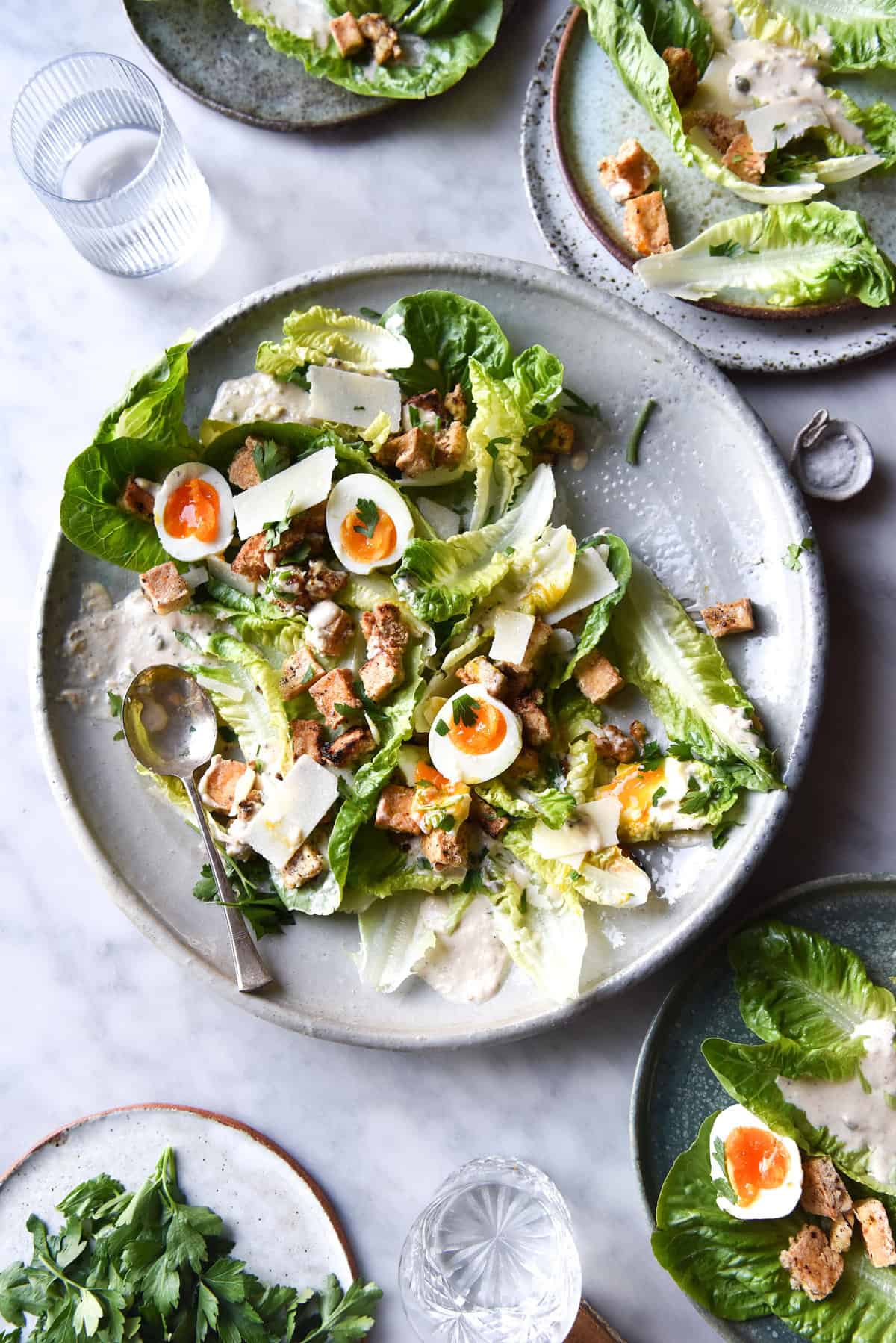 A vegetarian Caesar salad atop a large white ceramic serving platter. Other plates of Caesar salad and glasses of water surround the central serving platter which sits on a white marble table.