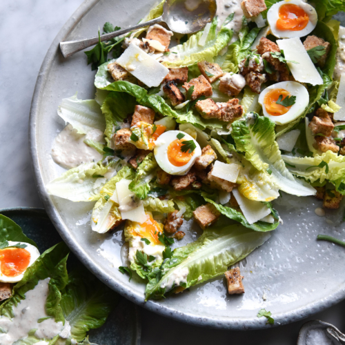 A vegetarian Caesar salad atop a large white ceramic serving platter. Other plates of Caesar salad and glasses of water surround the central serving platter which sits on a white marble table.