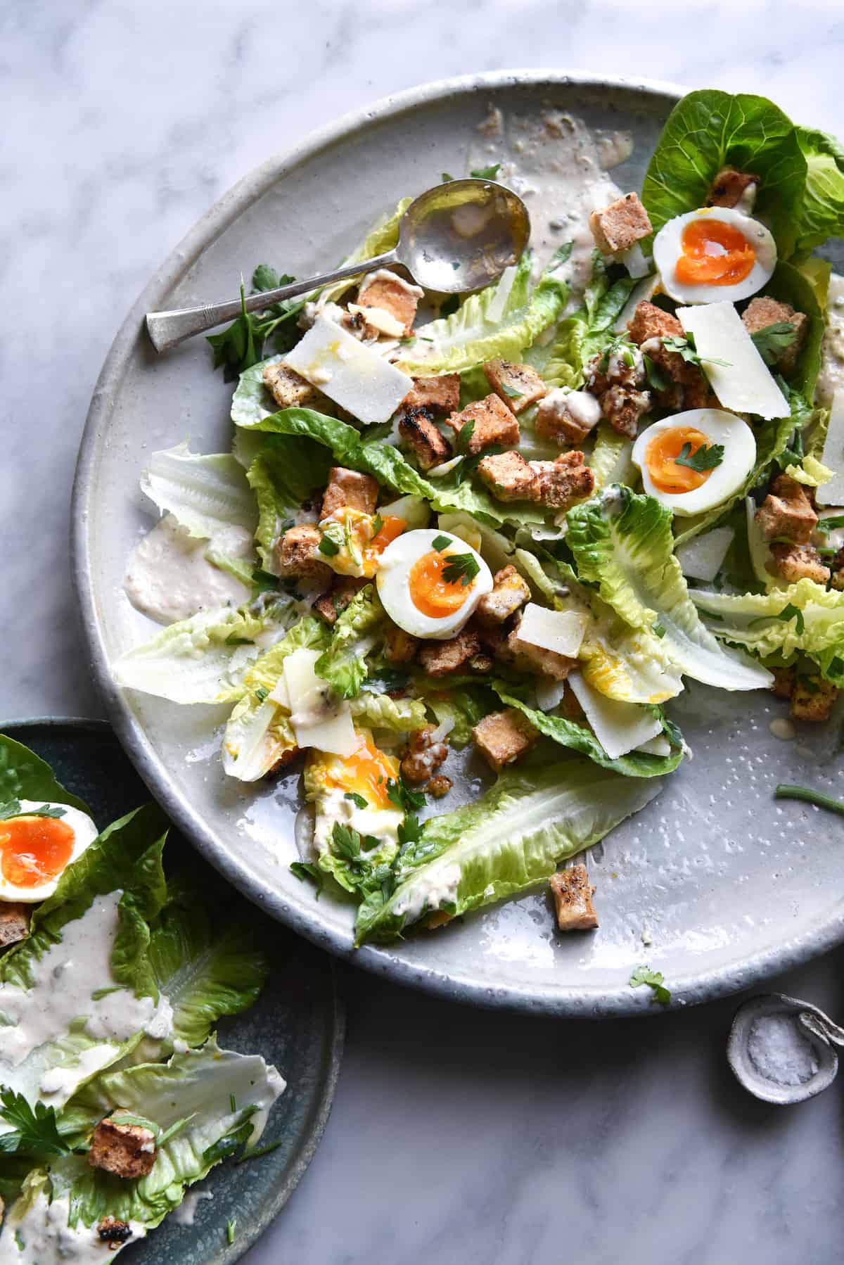 A vegetarian Caesar salad atop a large white ceramic serving platter. Other plates of Caesar salad and glasses of water surround the central serving platter which sits on a white marble table.