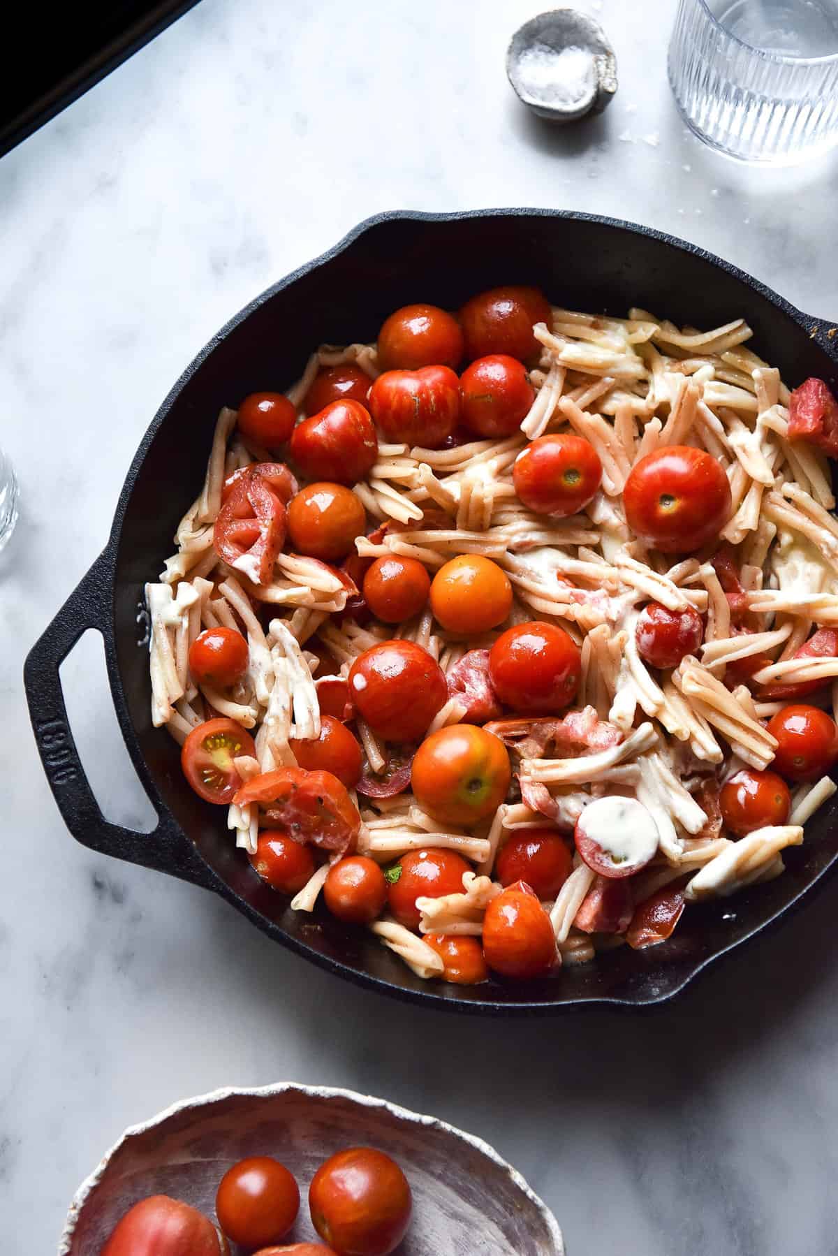 Summer tomato, lemon and mascarpone pasta in a skillet on a white marble table. The skillet is surrounded by water glasses and an extra bowl of pasta