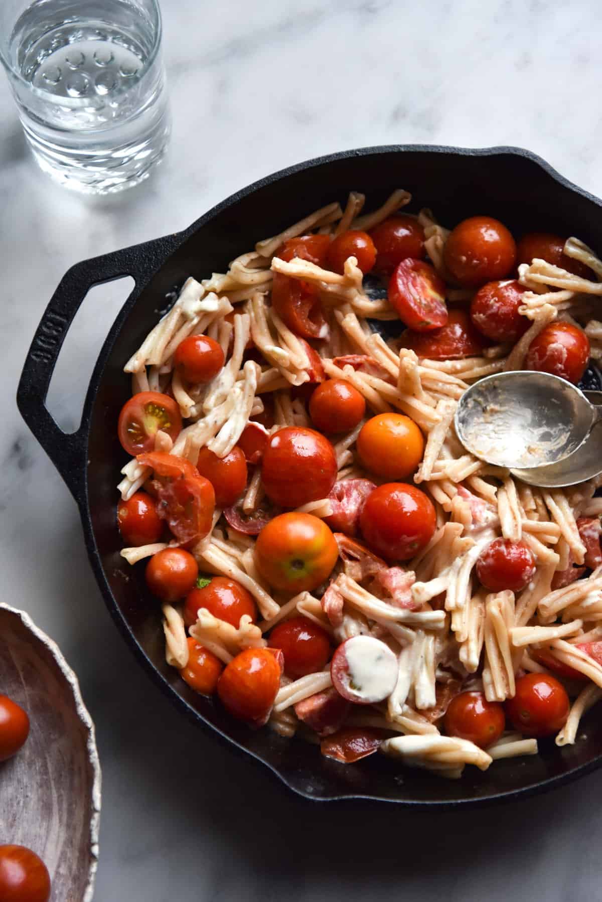 An aerial view of a skillet filled with summer tomato, lemon and mascarpone pasta. The skillet sits atop a white marble table and a bowl of extra tomatoes sits to the bottom left of the image. 