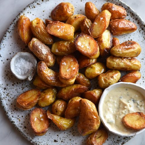 Vegan Caesar salad dressing in a white ceramic bowl on a plate topped with crispy roast potatoes. The plate sits atop a white marble table.