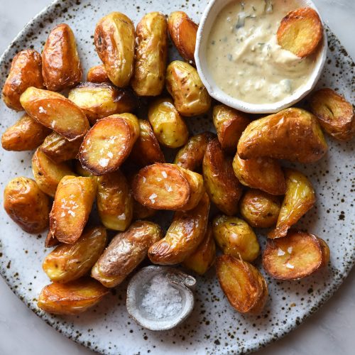 An aerial close up image of crispy Kipfler potatoes on a white speckled ceramic plate atop a white marble table. A small ceramic bowl of vegan caesar salad dressing sits in the top right corner of the plate of potatoes, and a small pinch bowl of salt sits in the bottom left corner.