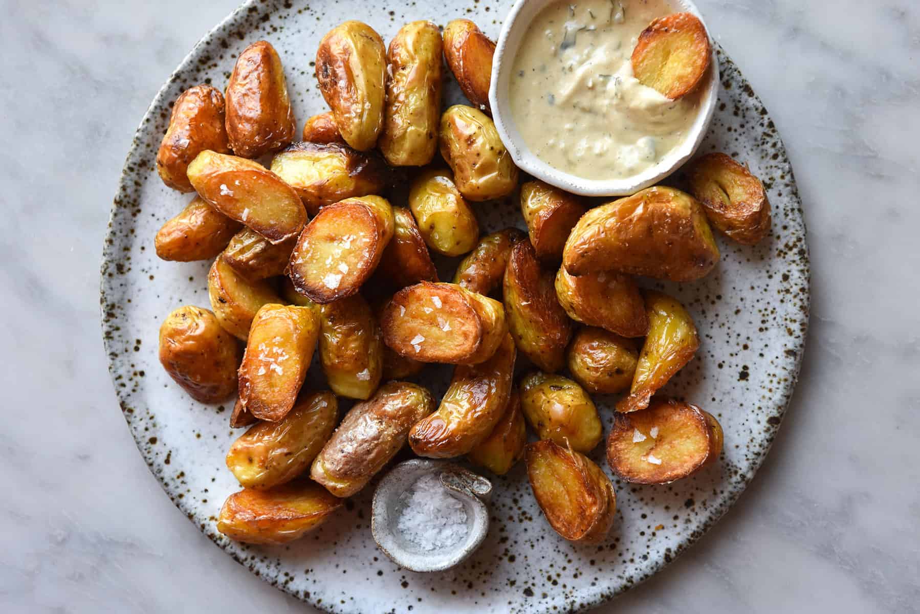 An aerial close up image of crispy Kipfler potatoes on a white speckled ceramic plate atop a white marble table. A small ceramic bowl of vegan caesar salad dressing sits in the top right corner of the plate of potatoes, and a small pinch bowl of salt sits in the bottom left corner. 