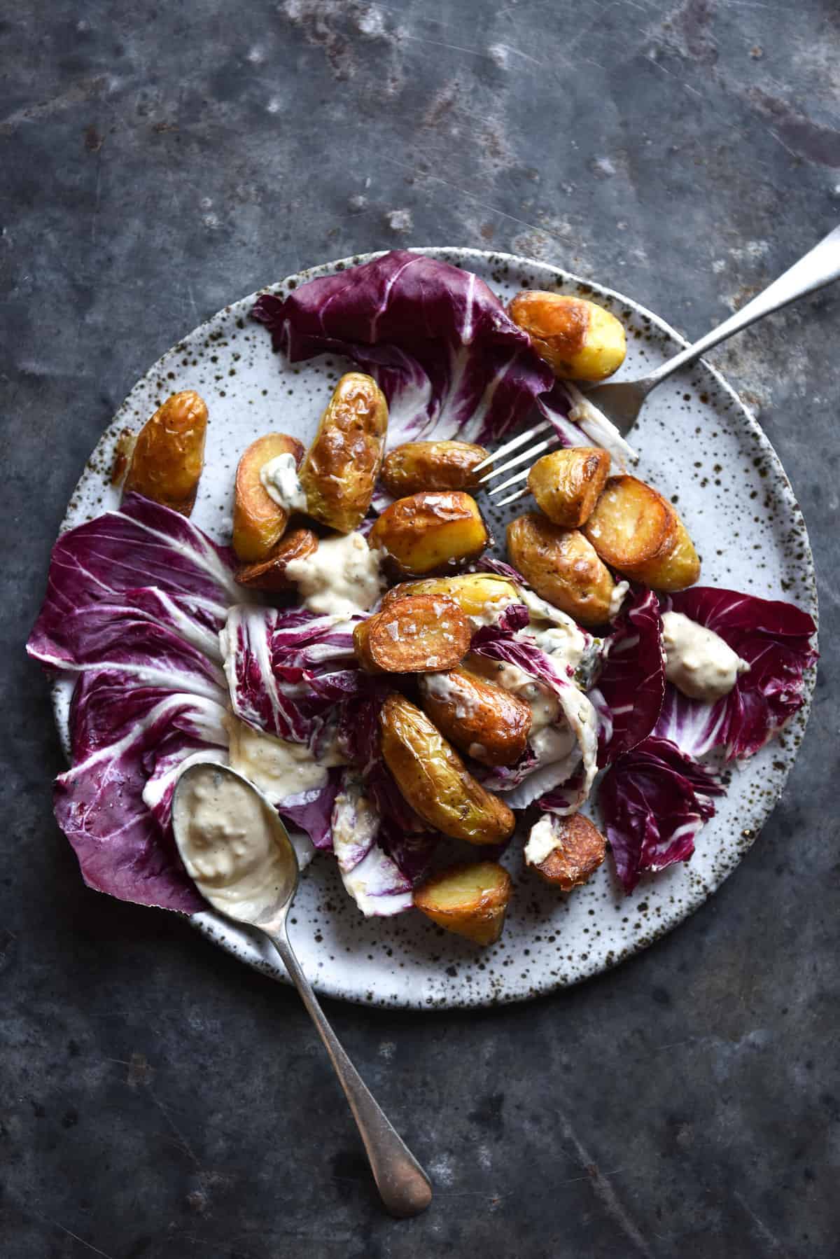 An aerial image of a plate of radicchio, potatoes and salad on a mottled dark blue steel backdrop.