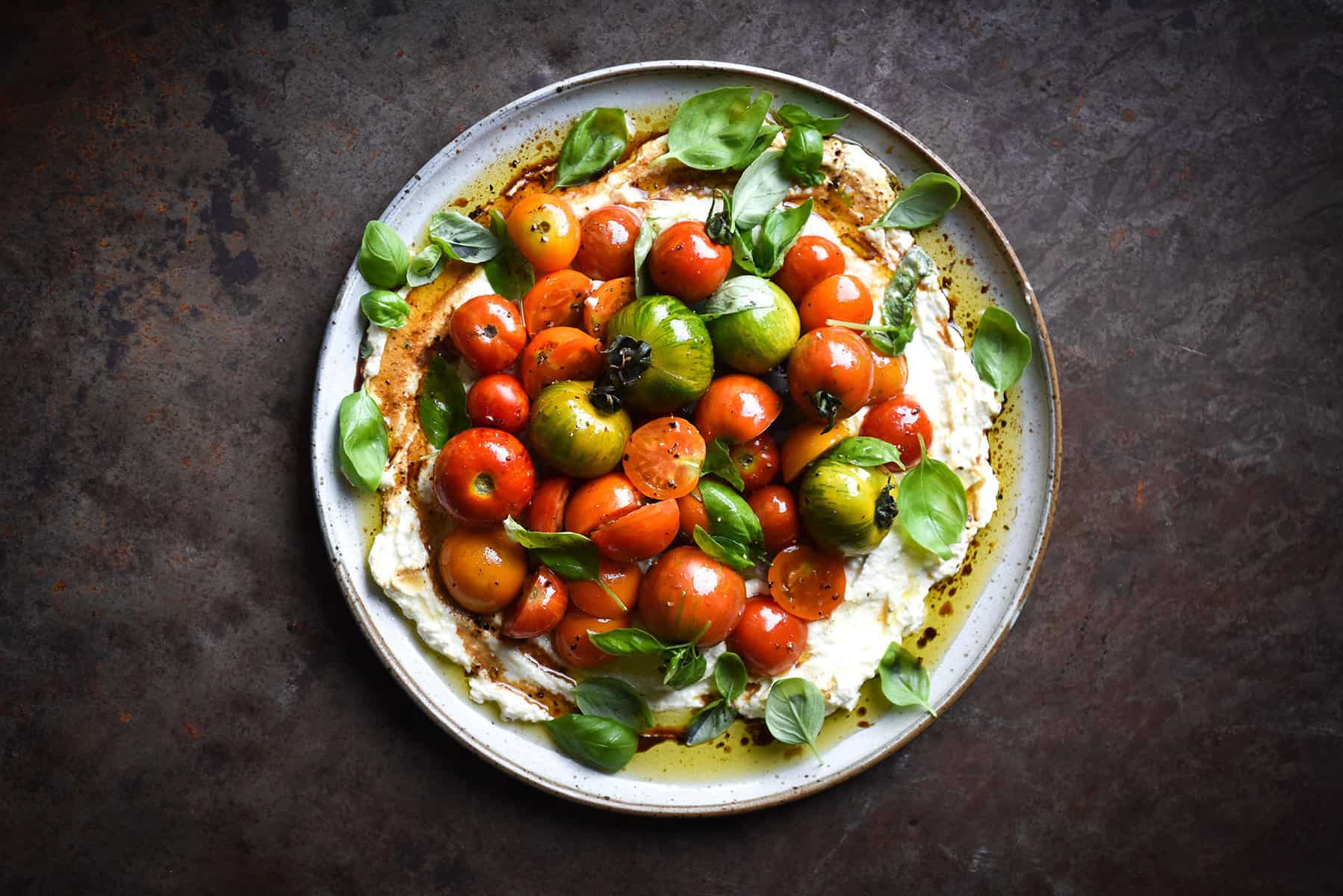 An aerial image of a white ceramic plate of ricotta and heirloom tomatoes on a mottled grey backdrop. 