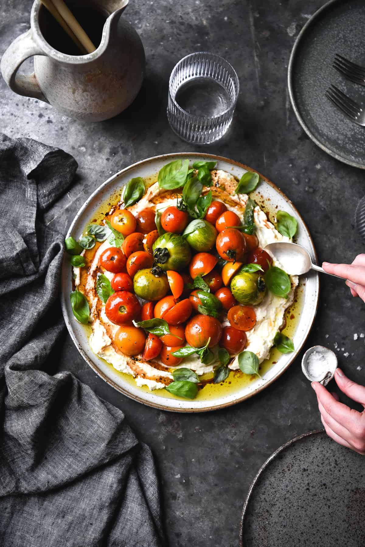 An aerial view of a plate of summer tomatoes on whipped lactose free ricotta. The dish is set on a ceramic white plate and the multicoloured summer tomatoes contrast with basil leaves. The scene is set on a blue backdrop surrounded by blue toned plates and tea towels. A hand extends from the right of the image to scoop some tomatoes up. The image is part of a guide to Vegetarian Cheese in Australia.