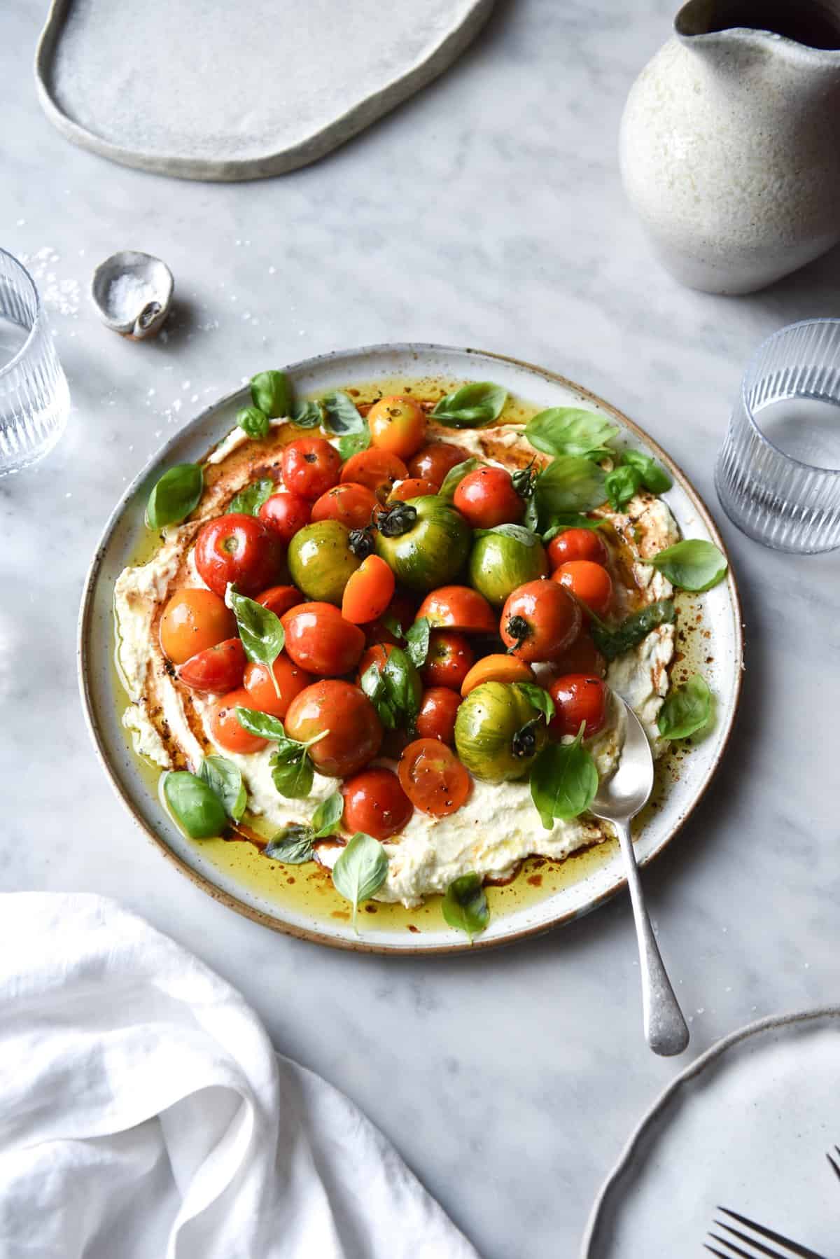 An aerial view of a plate of lactose-free lemon ricotta with summer tomatoes, balsamic and basil. The salad sits atop a white ceramic serving dish on a white marble table. The dish is surrounded by white linen, plates and crockery, which contrasts with the beautiful vibrance of the tomatoes.