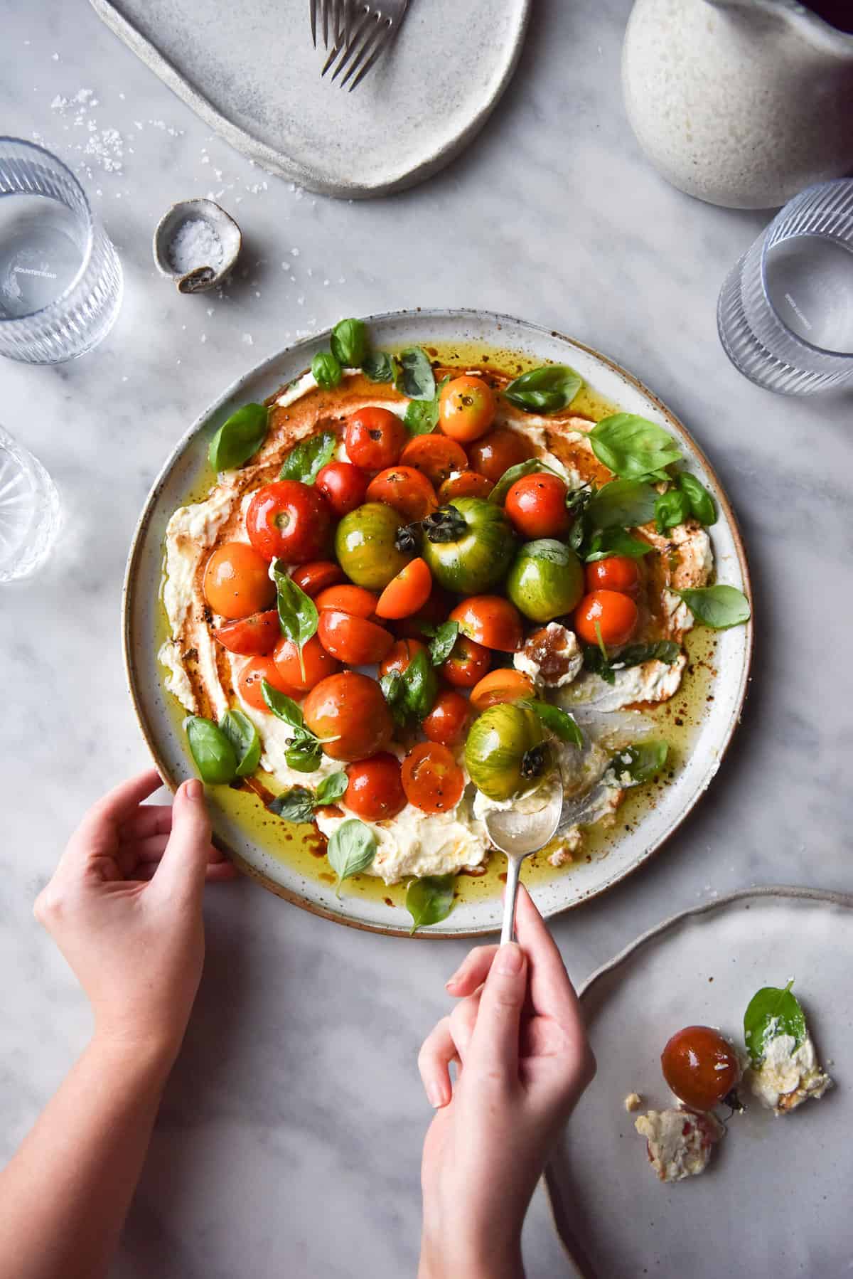 Lactose-free lemon ricotta with summer tomatoes, balsamic and basil on a white ceramic plate atop a white marble table. The salad is surrounded by water glasses, pinch bowls and extra white ceramics