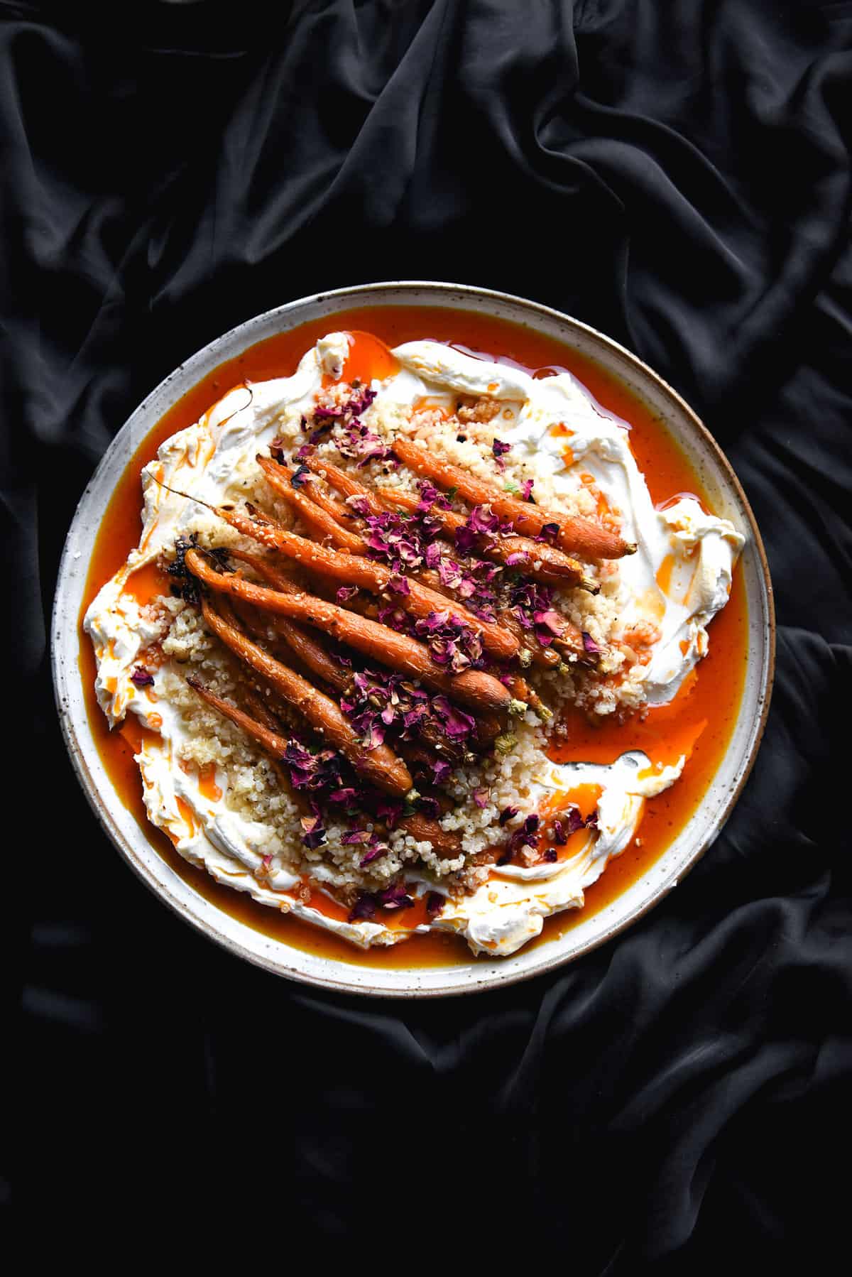 Honey and dukkah roasted carrots with lactose free labne, lemon rosewater quinoa and Aleppo chilli oil on a white ceramic plate atop a dark silky backdrop