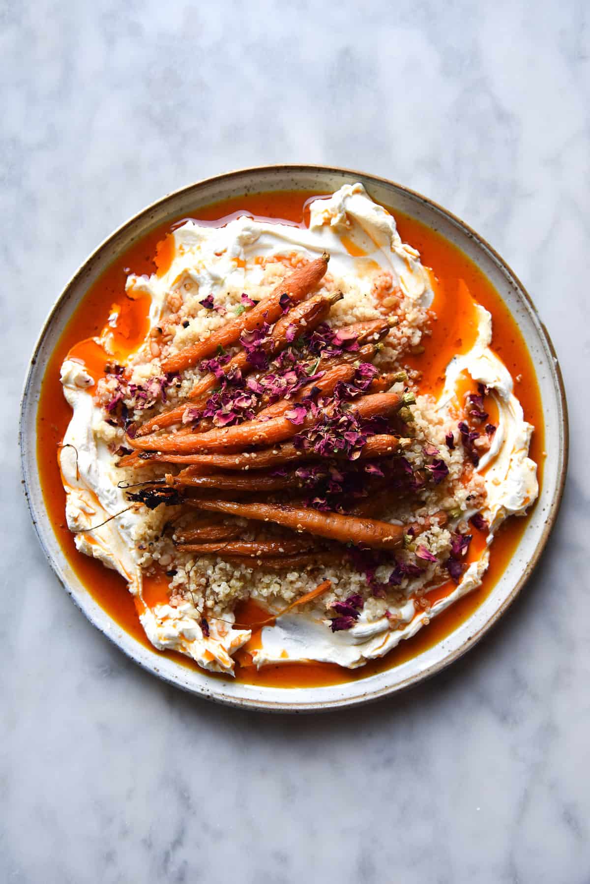 An aerial image of honey and dukkah roasted carrots with homemade lactose free labne, Aleppo chilli oil and lemon quinoa. The dish sits on a white ceramic plate atop a white marble table. 