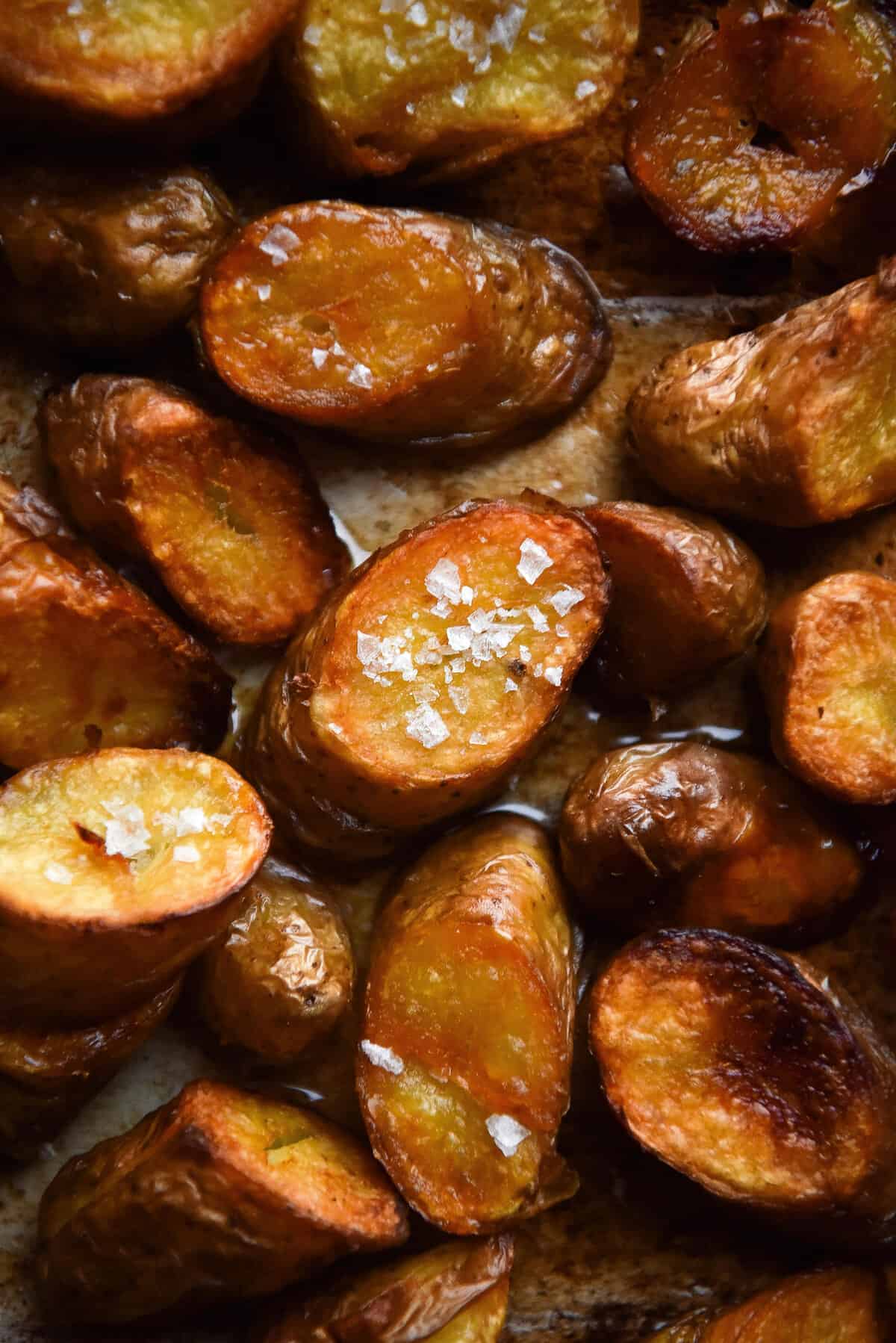 A close up moody image of crispy golden brown Kipfler potatoes on a baking dish. The central potato faces upwards and is covered in sea salt flakes. 