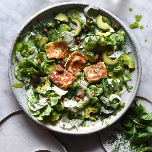 An aerial sunlit view of a green salad topped with haloumi. The salad sits on a white plate against a white marble table