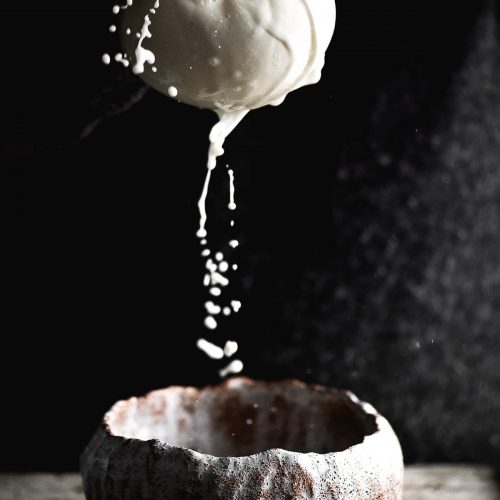 A side on moody photo of a hand holding out a muslin cloth filled with lactose free ricotta and squeezing out the liquid. The liquid falls into a ceramic bowl at the bottom of the image, and the white droplets contrast dramatically against the dark backdrop