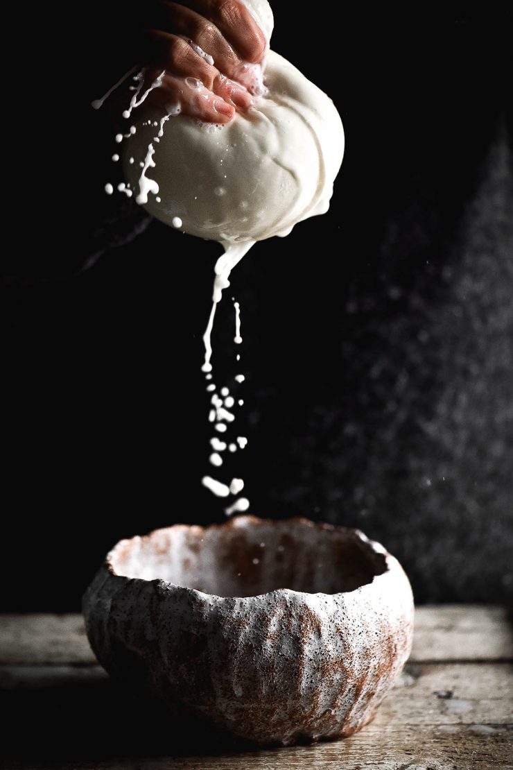 A side on moody photo of a hand holding out a muslin cloth filled with lactose free ricotta and squeezing out the liquid. The liquid falls into a ceramic bowl at the bottom of the image, and the white droplets contrast dramatically against the dark backdrop