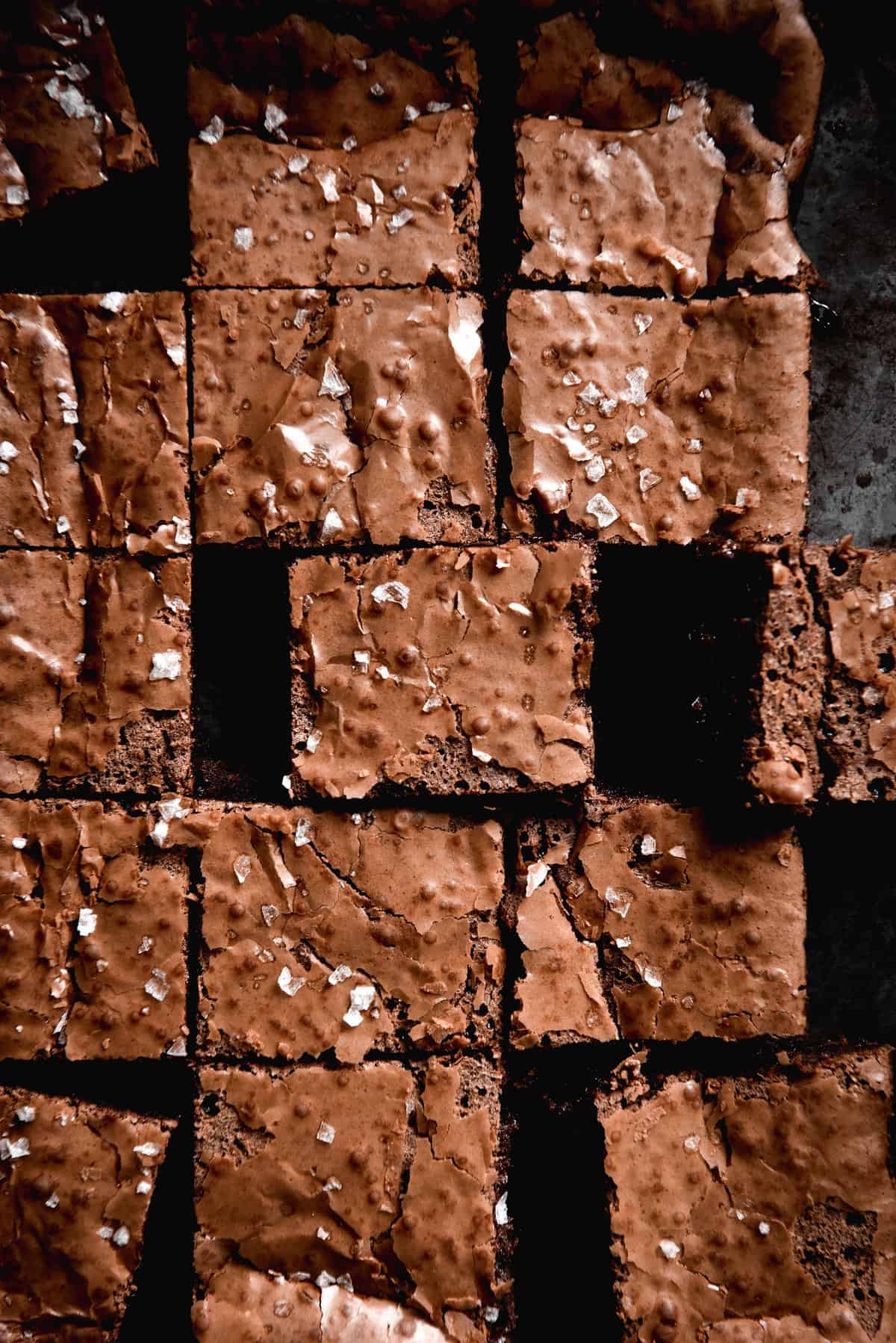 An aerial moody shot of gluten free brownies sliced and arranged on a dark steel backdrop. The brownies have flaky tops and are finished with sea salt flakes