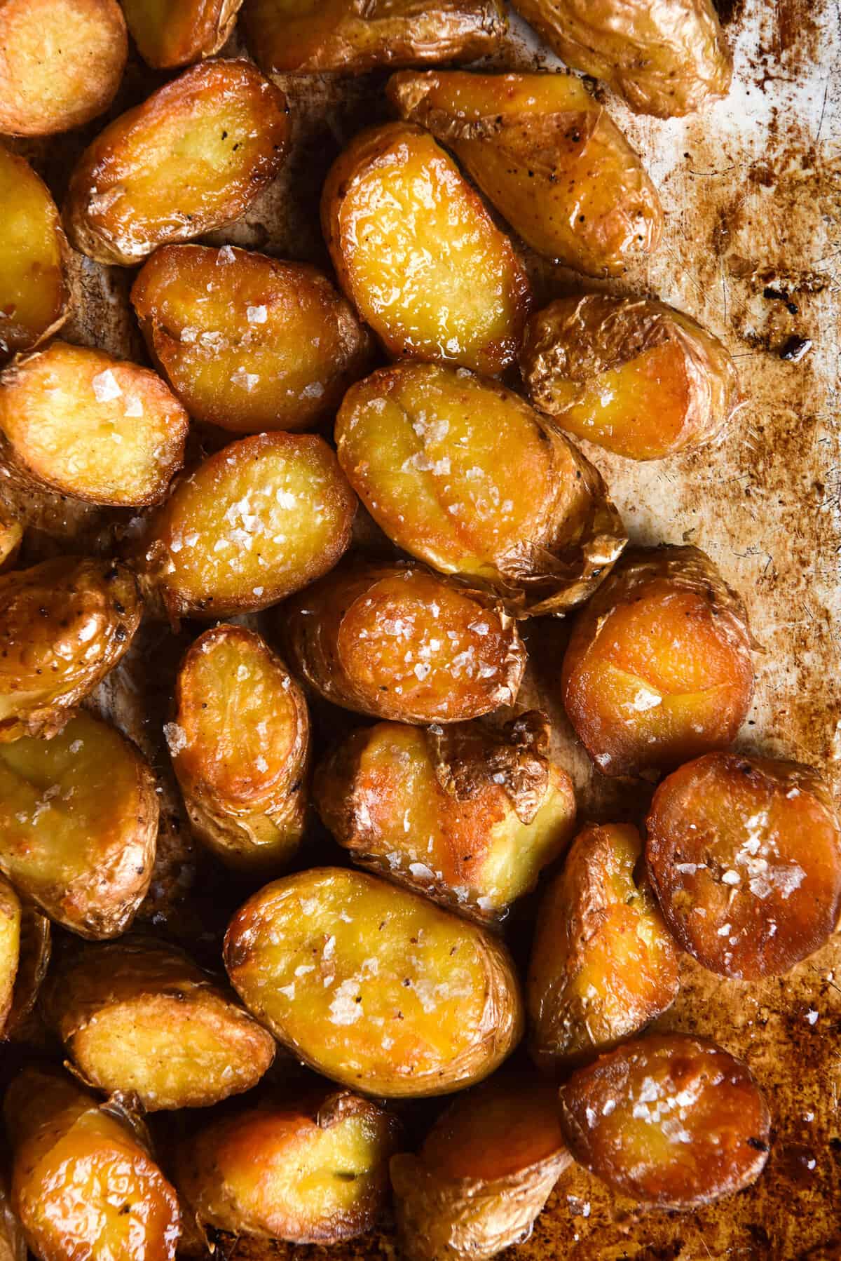 An aerial image of crispy golden brown kipfler potatoes on a mottled oven tray. The kipflers are topped with a sprinkle of sea salt flakes.