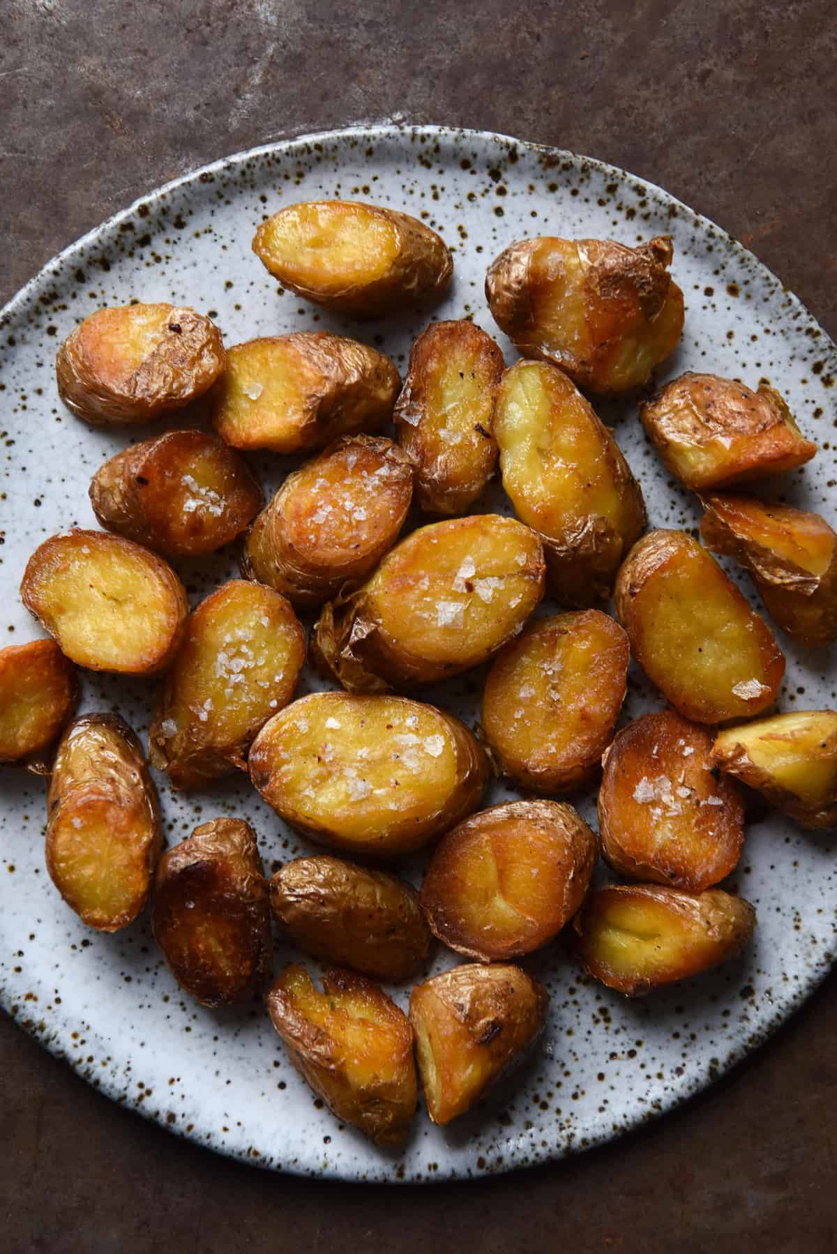 A close up aerial image of crispy, golden brown kipfler potatoes on a white speckled ceramic plate atop a mottled rust coloured backdrop. The potatoes are crispy and topped with flecks of sea salt flakes.
