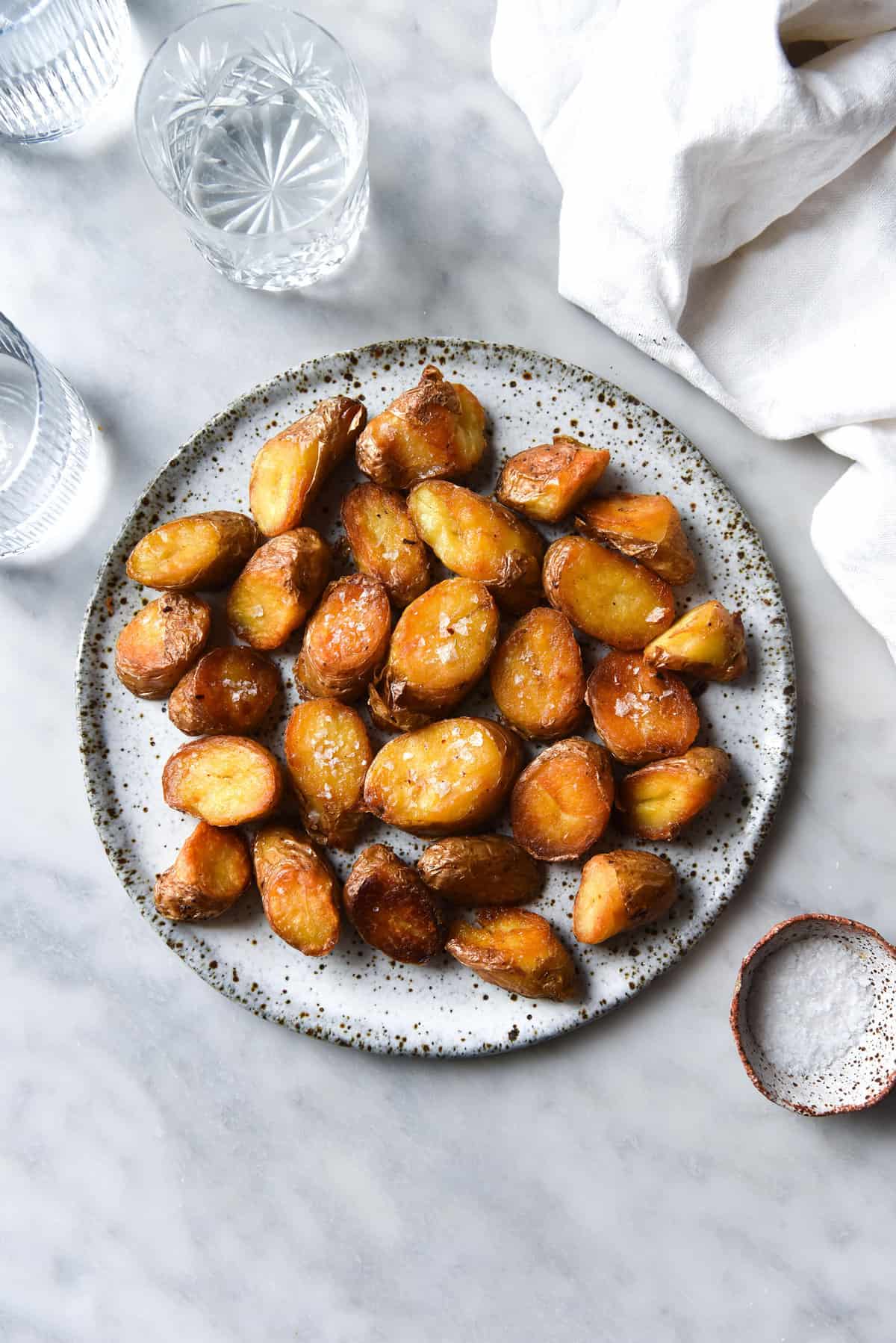 An aerial view of a white speckled ceramic plate topped with crispy roast potatoes. The potatoes are golden brown and topped with sea salt flakes. The plate sits on a white marble table surrounded by linen and pinch bowls of salt.