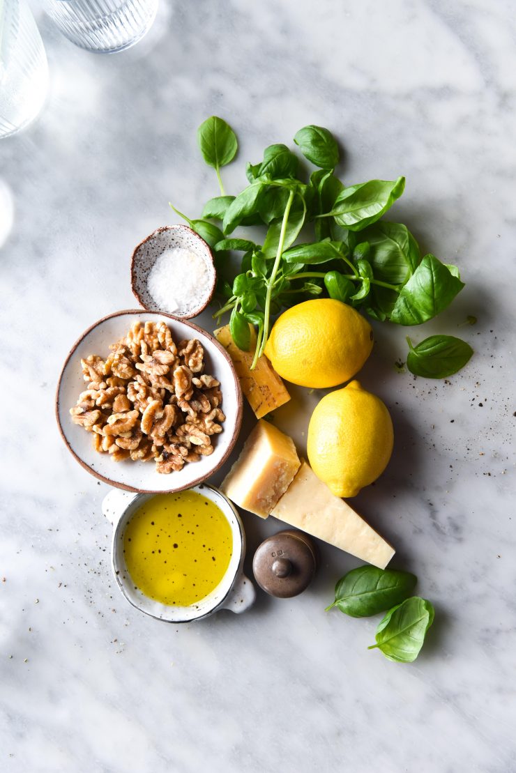 An aerial view of the ingredients for FODMAP friendly basil and walnut pesto arranged on a white marble table