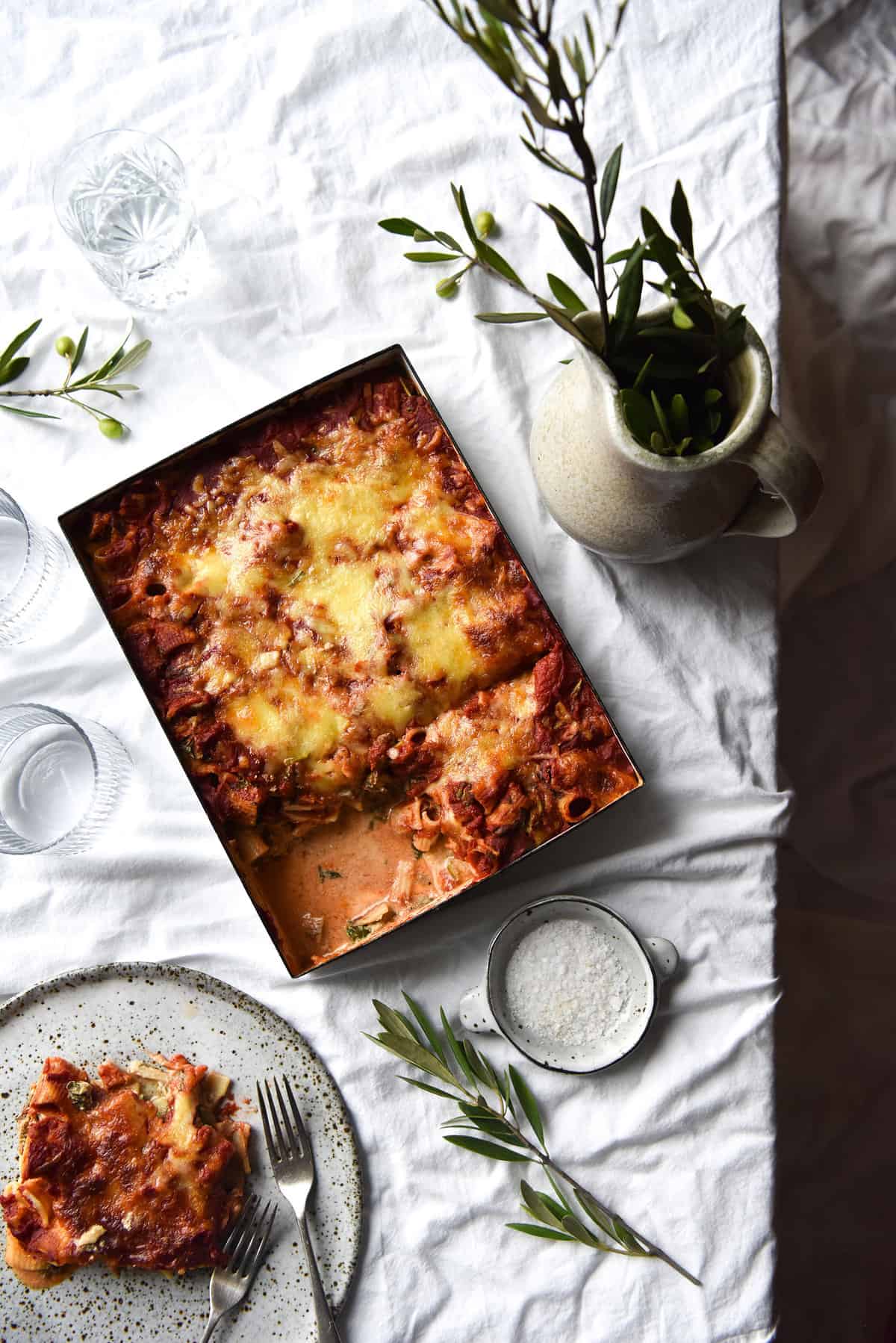 An aerial image of a gluten free pasta bake on a white marble table surrounded by olive leaves and an extra plate of pasta bake.