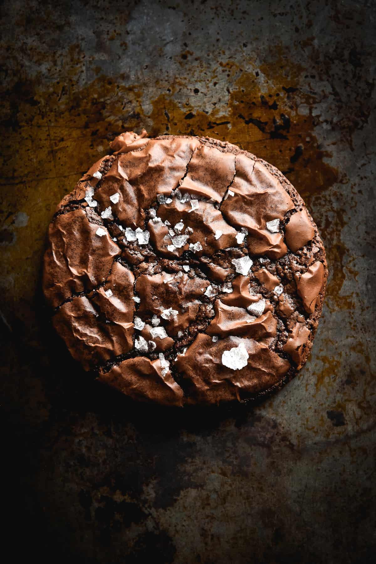 A moody aerial image of a gluten free brownie cookie that is crinkled on top and covered in sea salt flakes. The brownie sits atop a dark grey backdrop. 