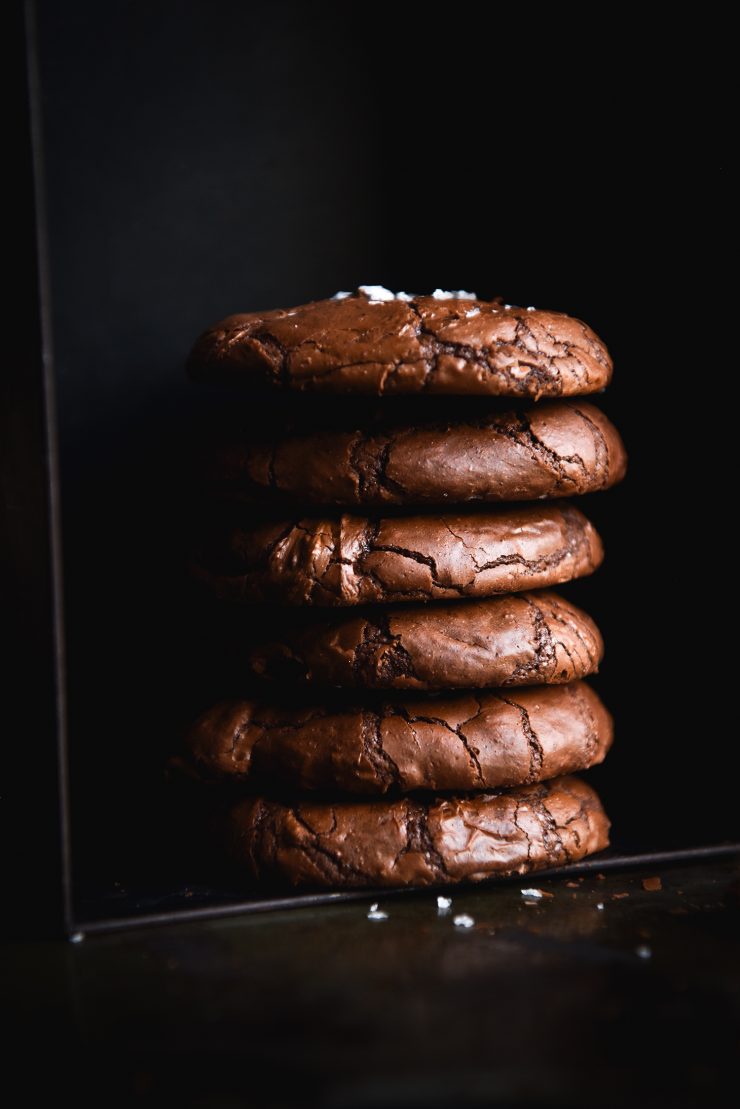A dark and moody side on image of a stack of gluten free brownie cookies against a black backdrop. The cookies are sprinkled with sea salt flakes which contrasts against the black backdrop.