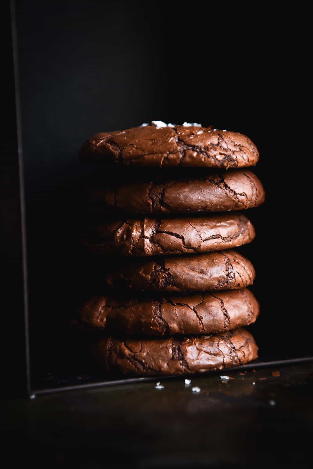 A dark and moody side on image of a stack of gluten free brownie cookies against a black backdrop. The cookies are sprinkled with sea salt flakes which contrasts against the black backdrop.